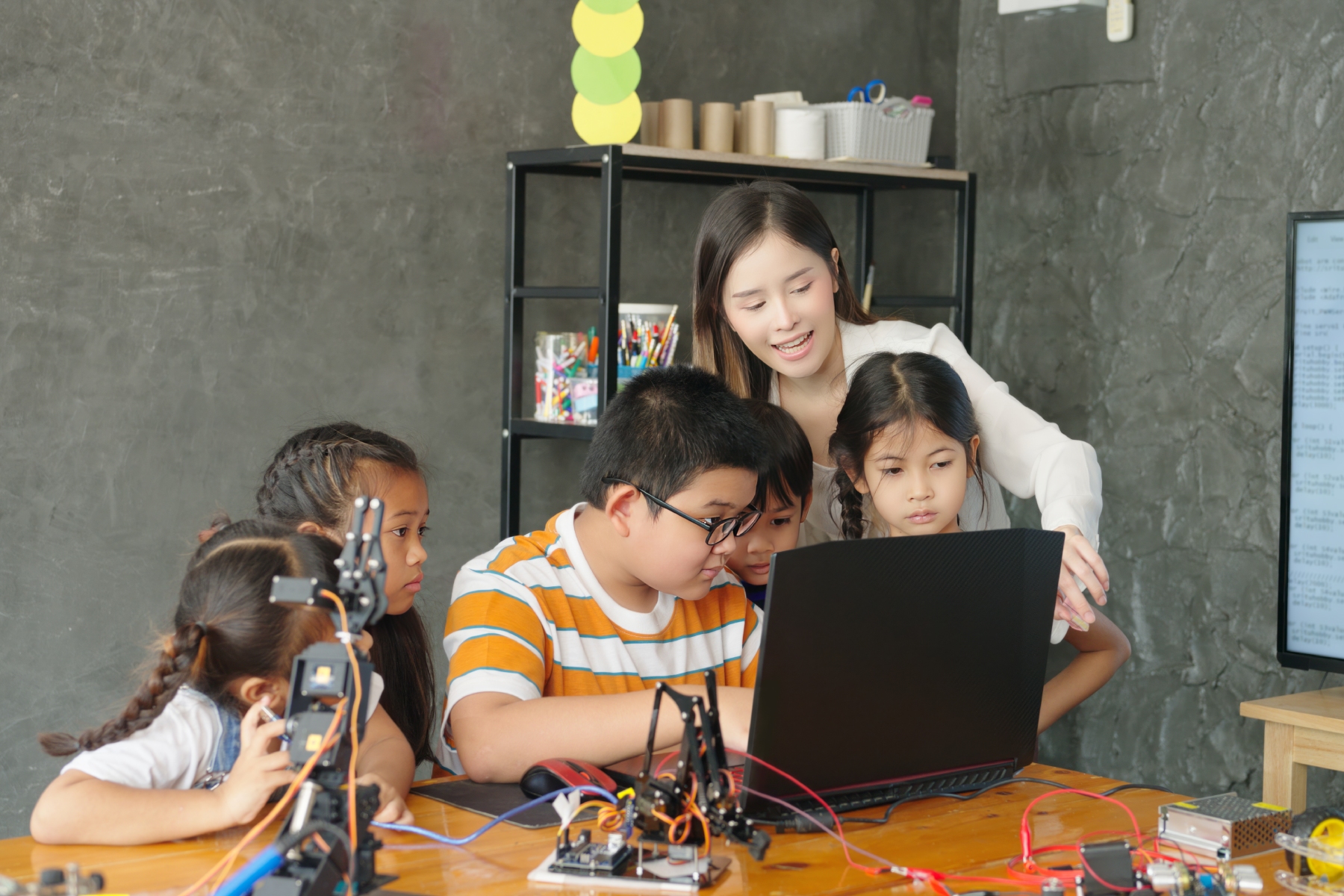 Teacher and students gather around a computer and a robot arm on a desk in a classroom