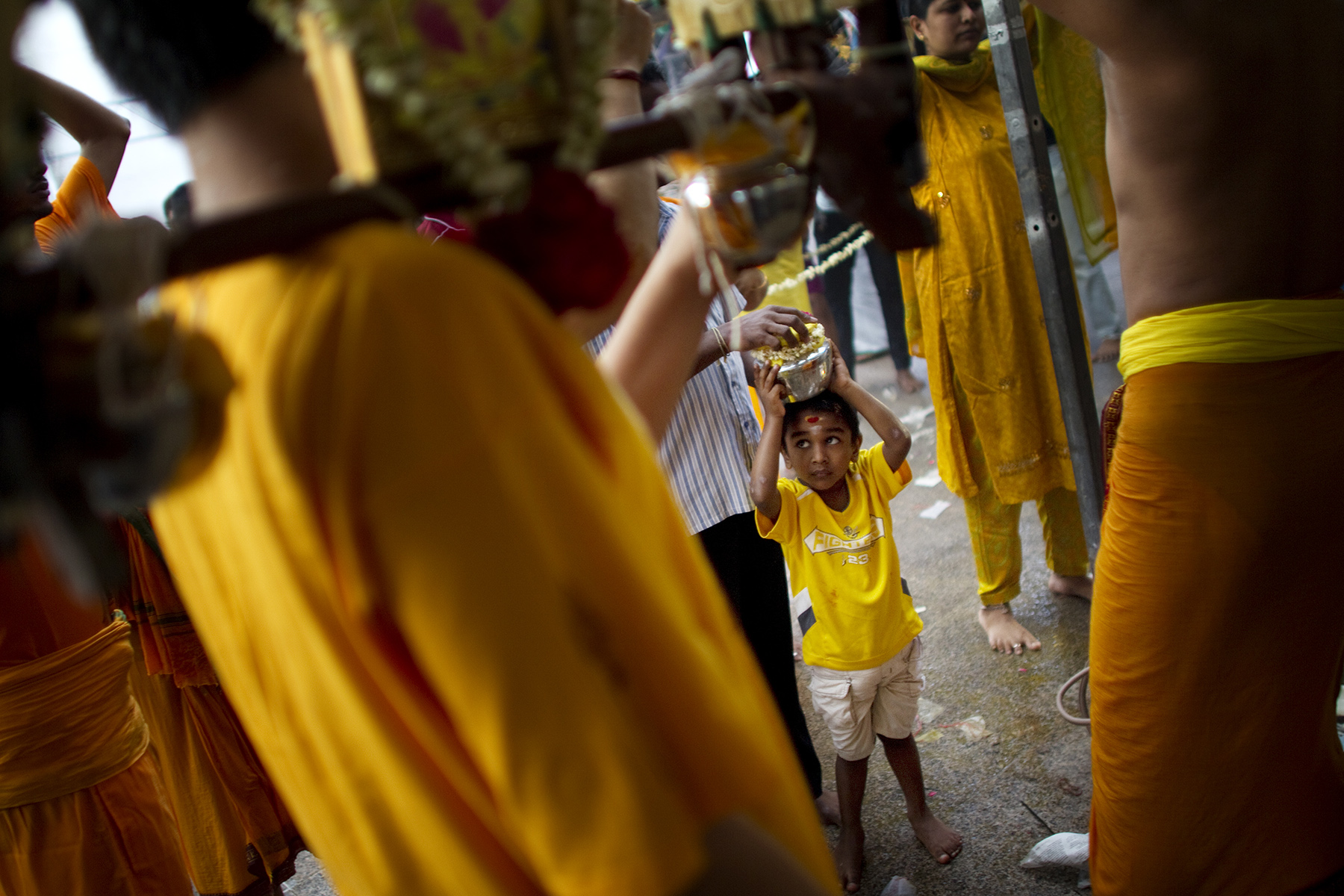 A young boy dressed in yellow, stands between adults, and holds a milk pot on his head before taking part in the Hindu Thaipusam procession 

