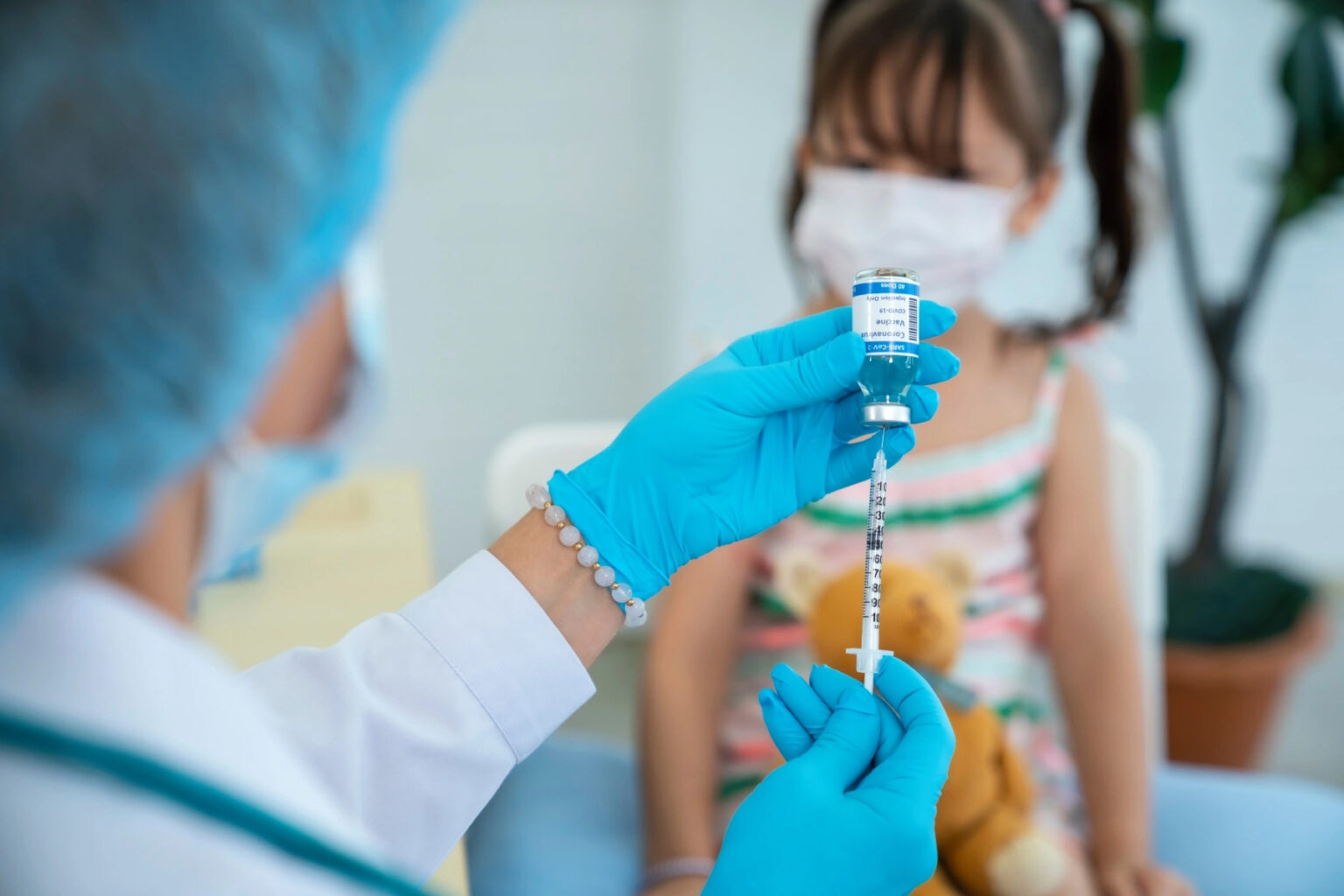 A doctor wearing scrubs prepared a vaccination in front of a young girl wearing colorful clothing and a face mask