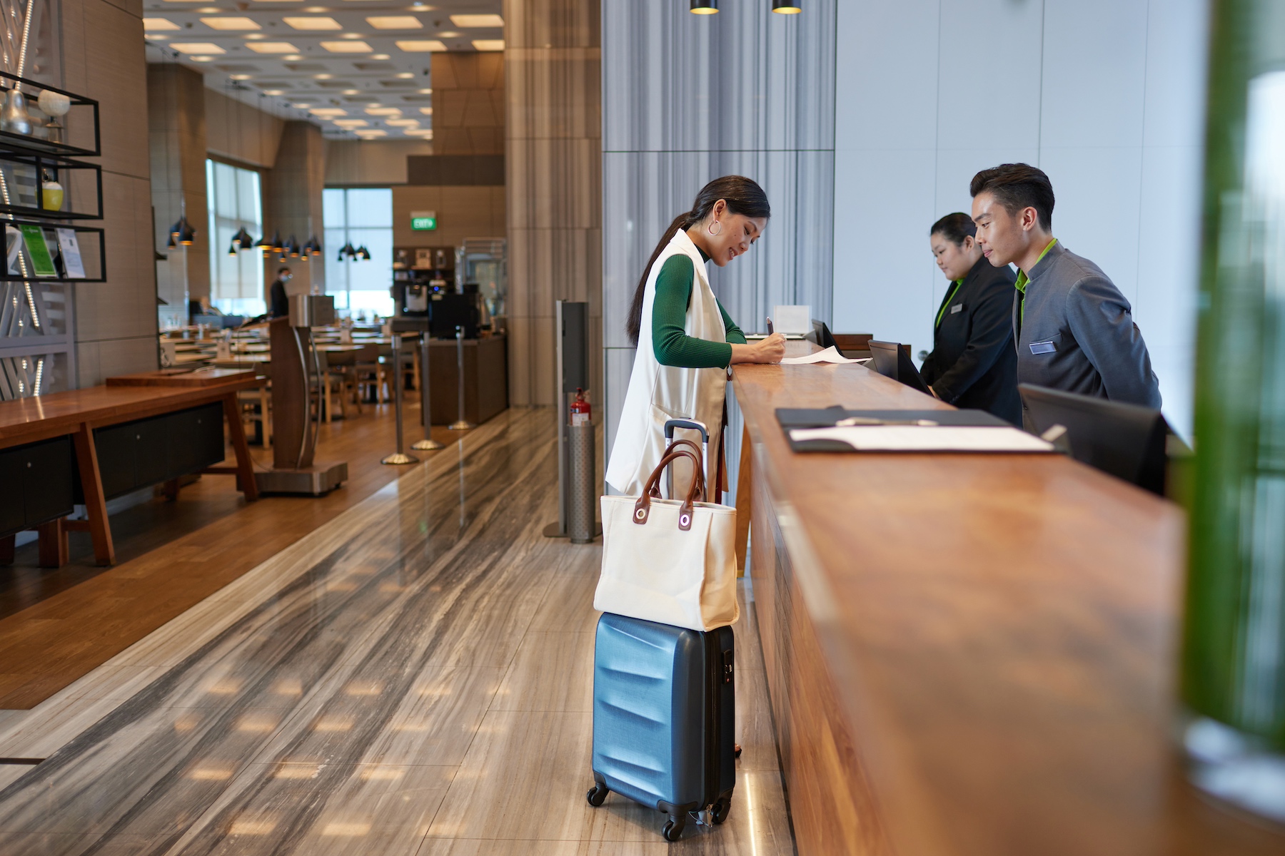 A woman stands at the front desk of a hotel with a suitcase