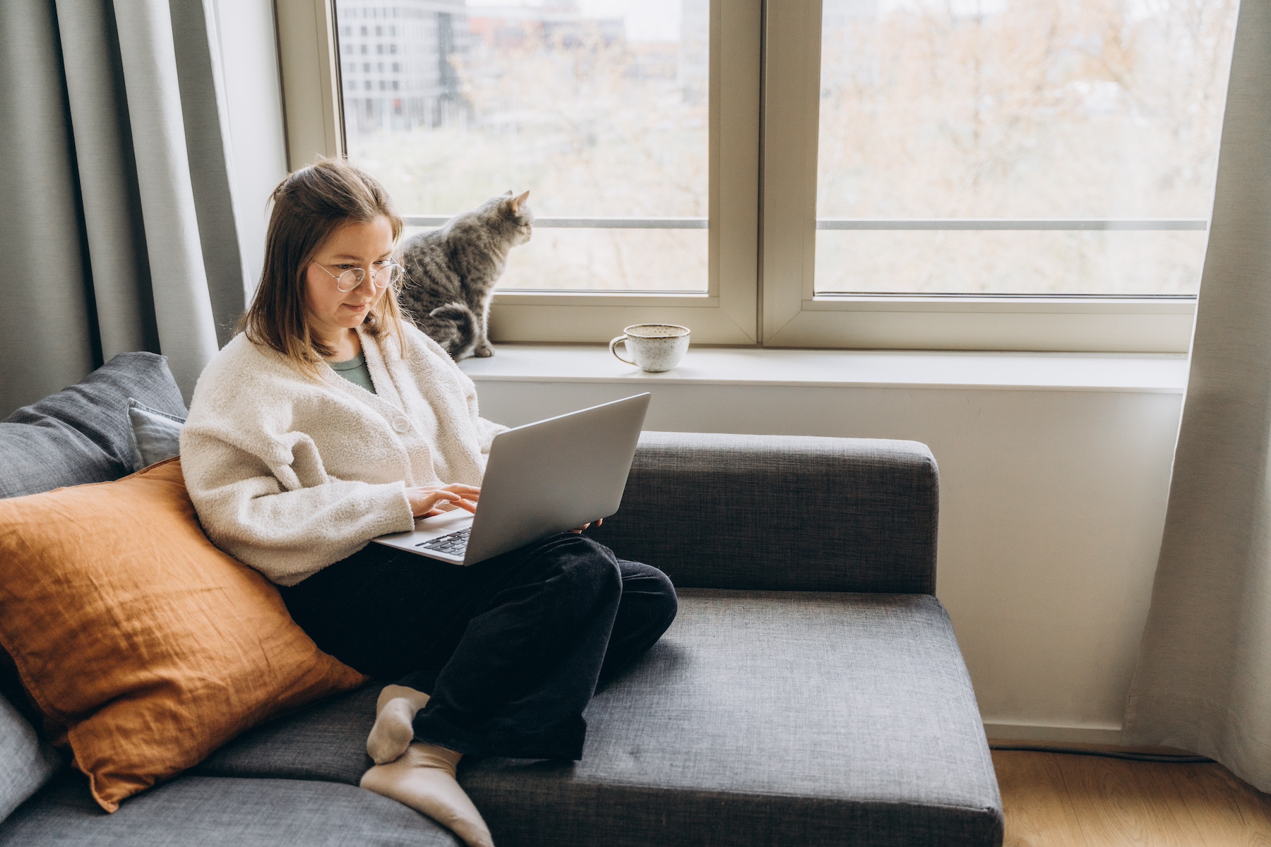A woman sits on the couch typing on a laptop with her cat behind her