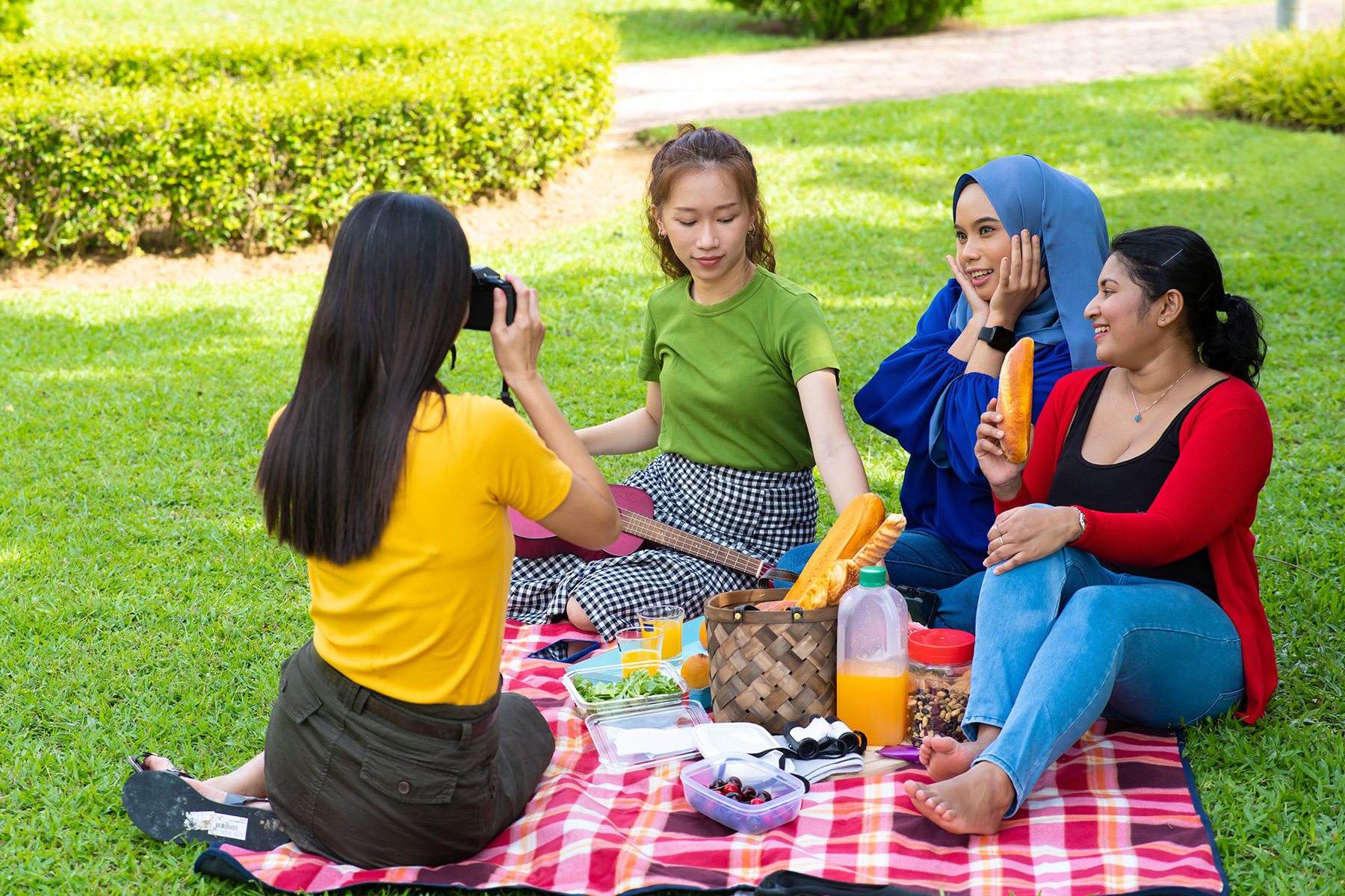 Women having a picnic in a park. One is posing for a picture, the others are focussed on the bread.