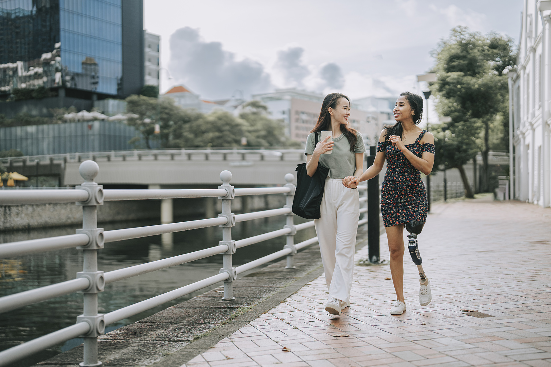 Two women holding hand s walks next to a river in Singapore. One has a prosthetic leg

