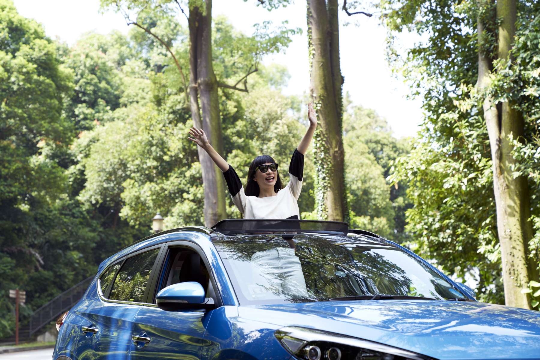 Woman with sunglasses cheering as she is standing through the opening of a sun roof of a blue car.