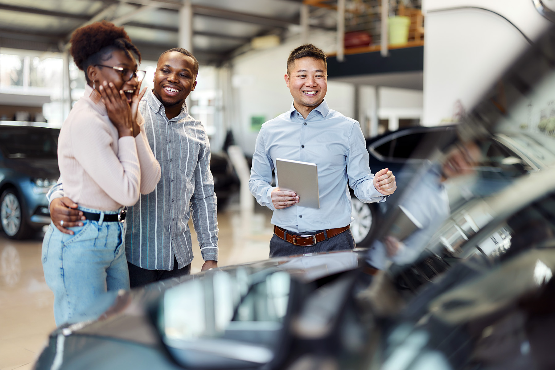 Couple is looking at a car inside a dealer's showroom. The lady has her hands on her face, smiling with her mouth open, the man and the salesperson are laughing giddily.