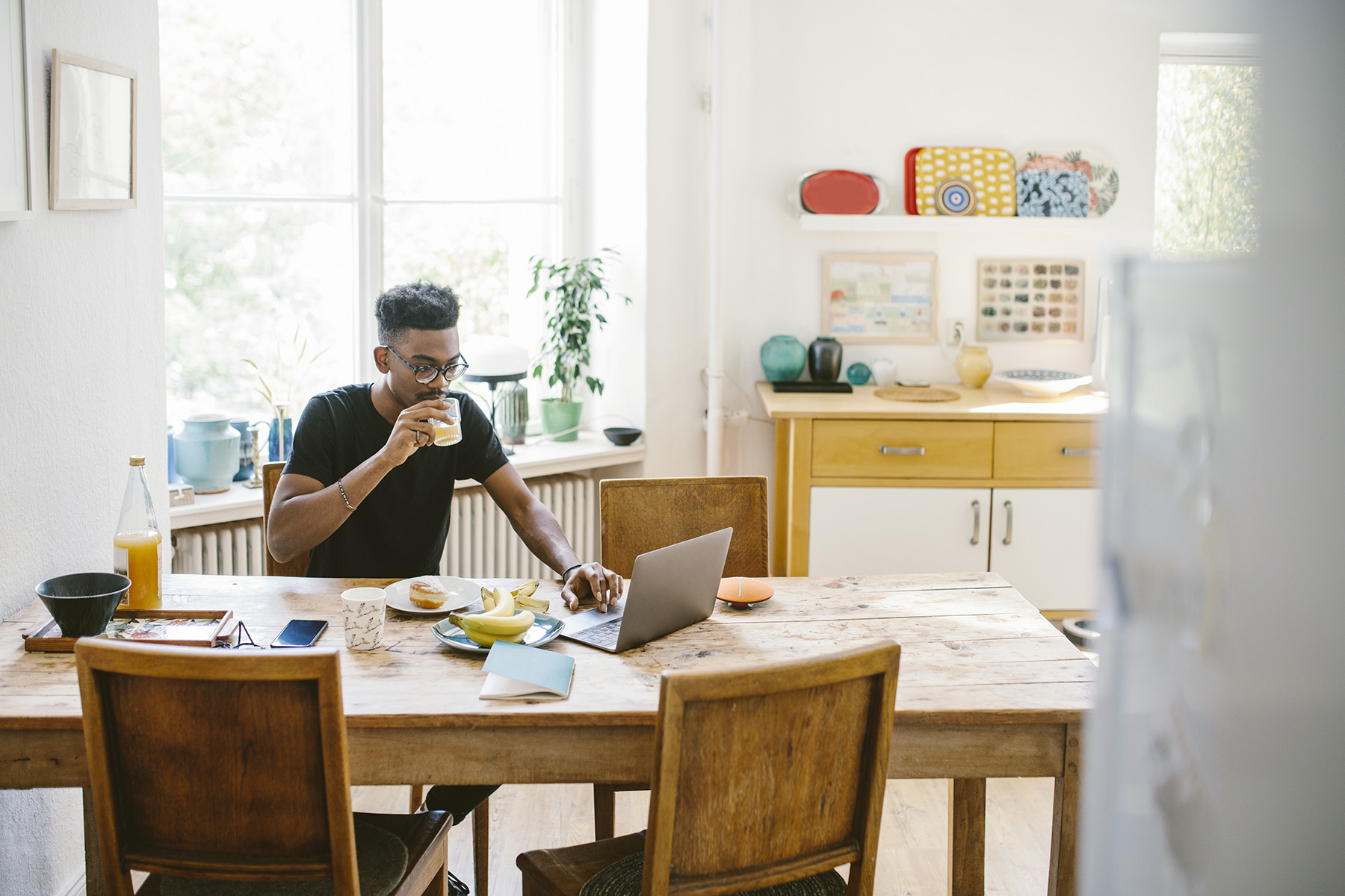 Man eating breakfast and drinking juice while he is working on a laptop at the kitchen table.
