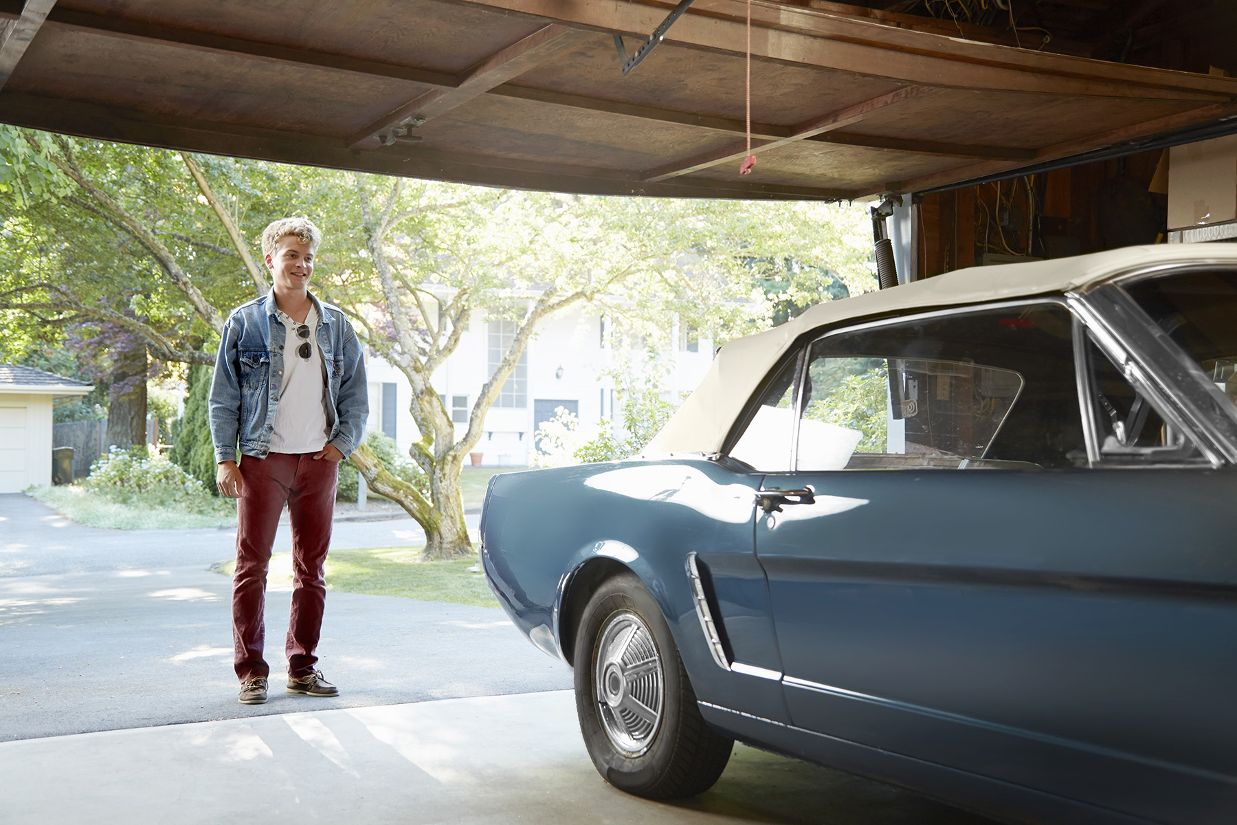 Young man happily staring at his vintage car inside a garage. He is dressed vintage too.
