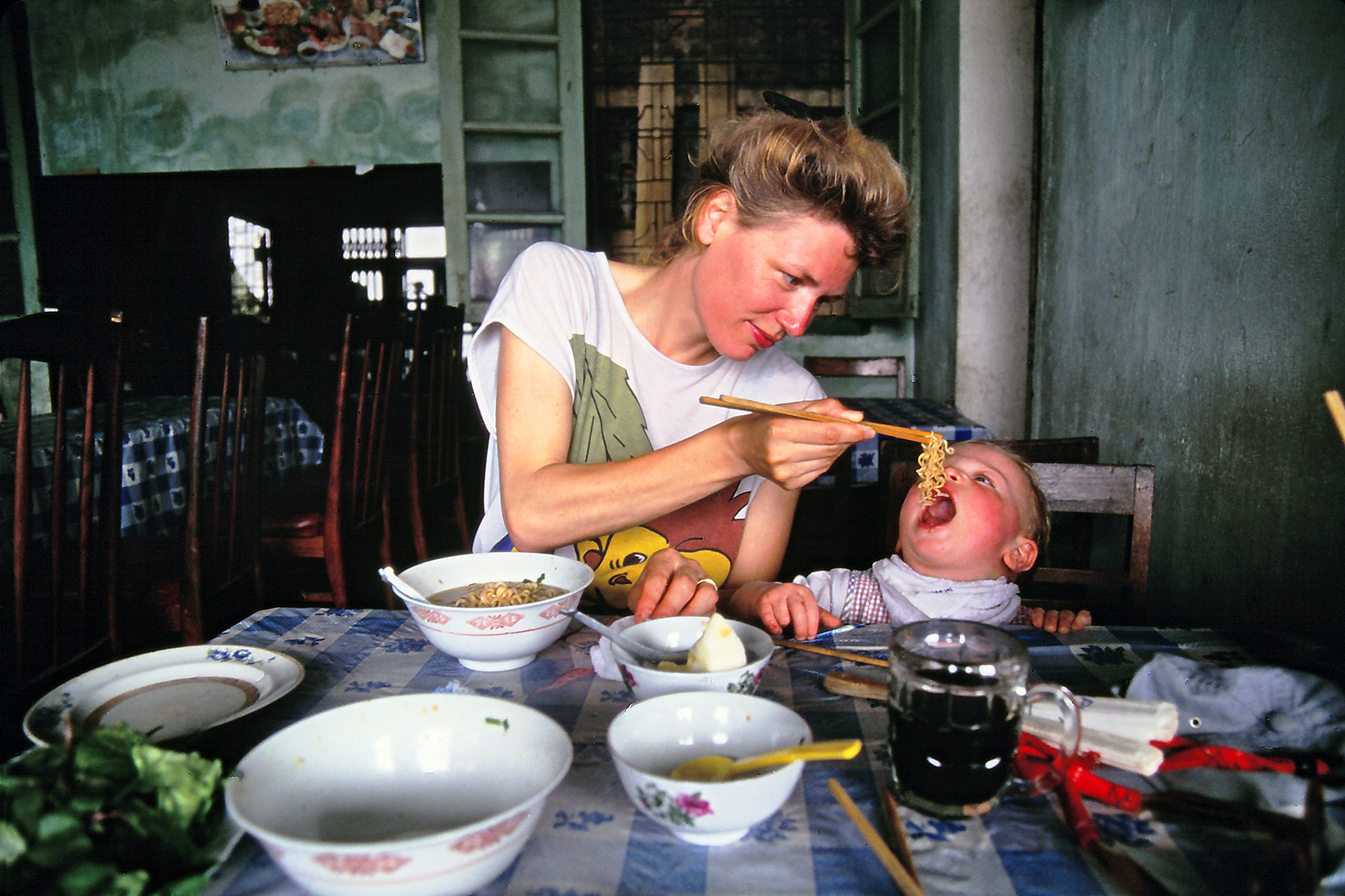 Mother bird-feeding her two year old son some noodles with chopsticks.