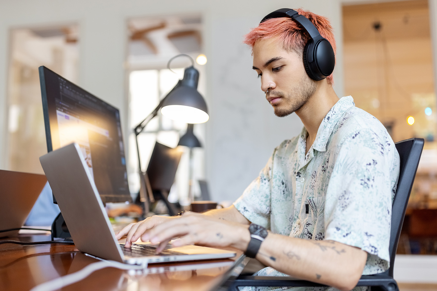 Man with pink hair and headphones working on laptop at an office.