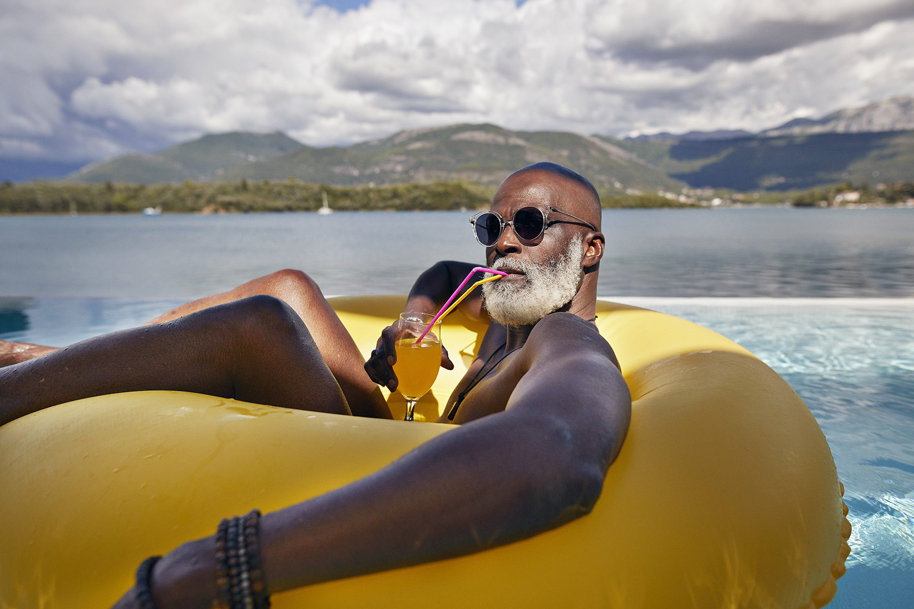 Bald man wearing sunglasses and drinking juice from a fancy glass, while chilling in a yellow inflatable ring in the pool during vacation.