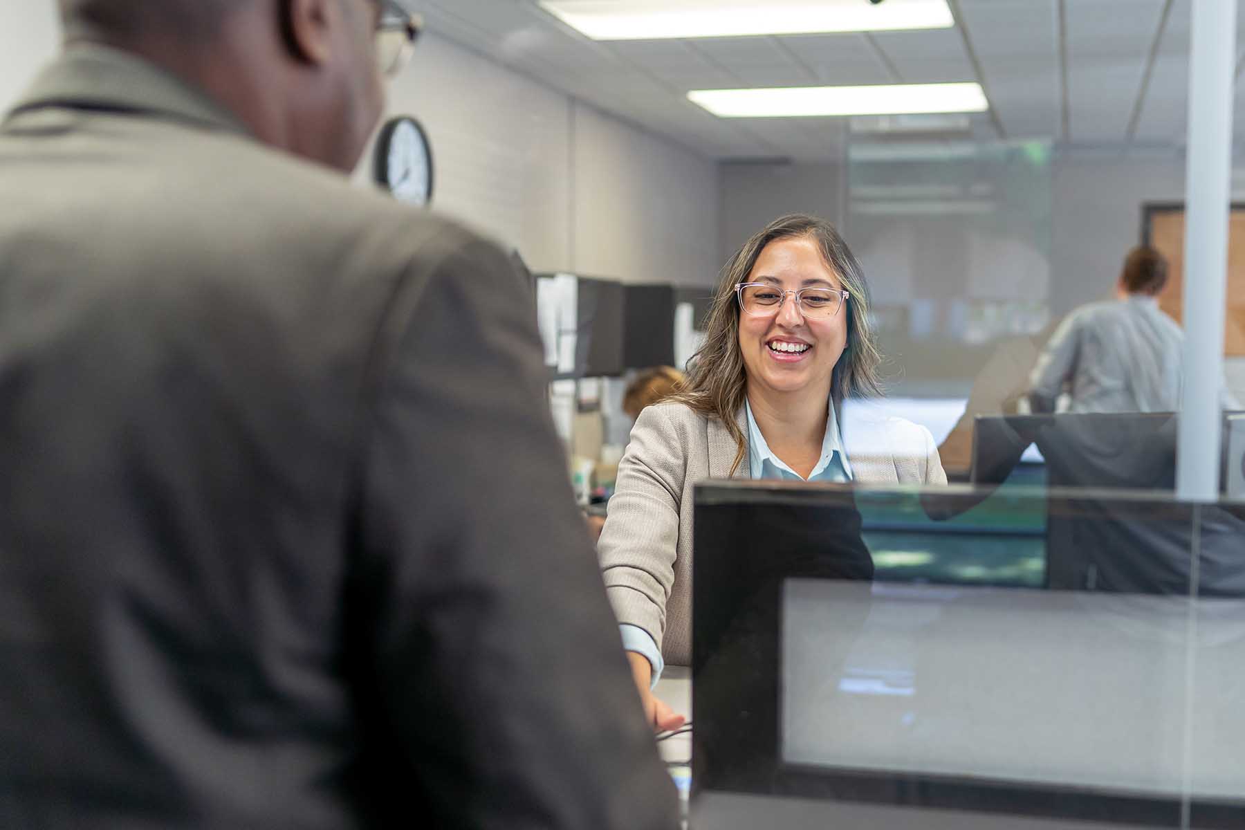 A female customer service representative of the bank smiles while she greet a customer behind the glass.