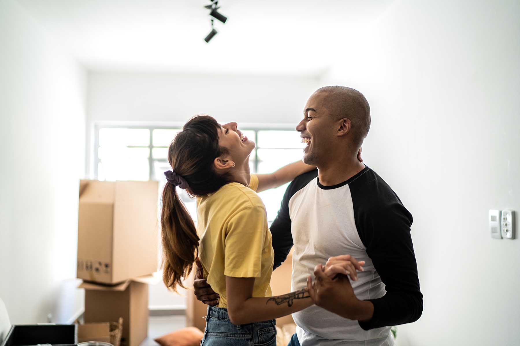 Couple laughing and dance-embracing in their new (empty) home. Moving boxes are visible in the background.