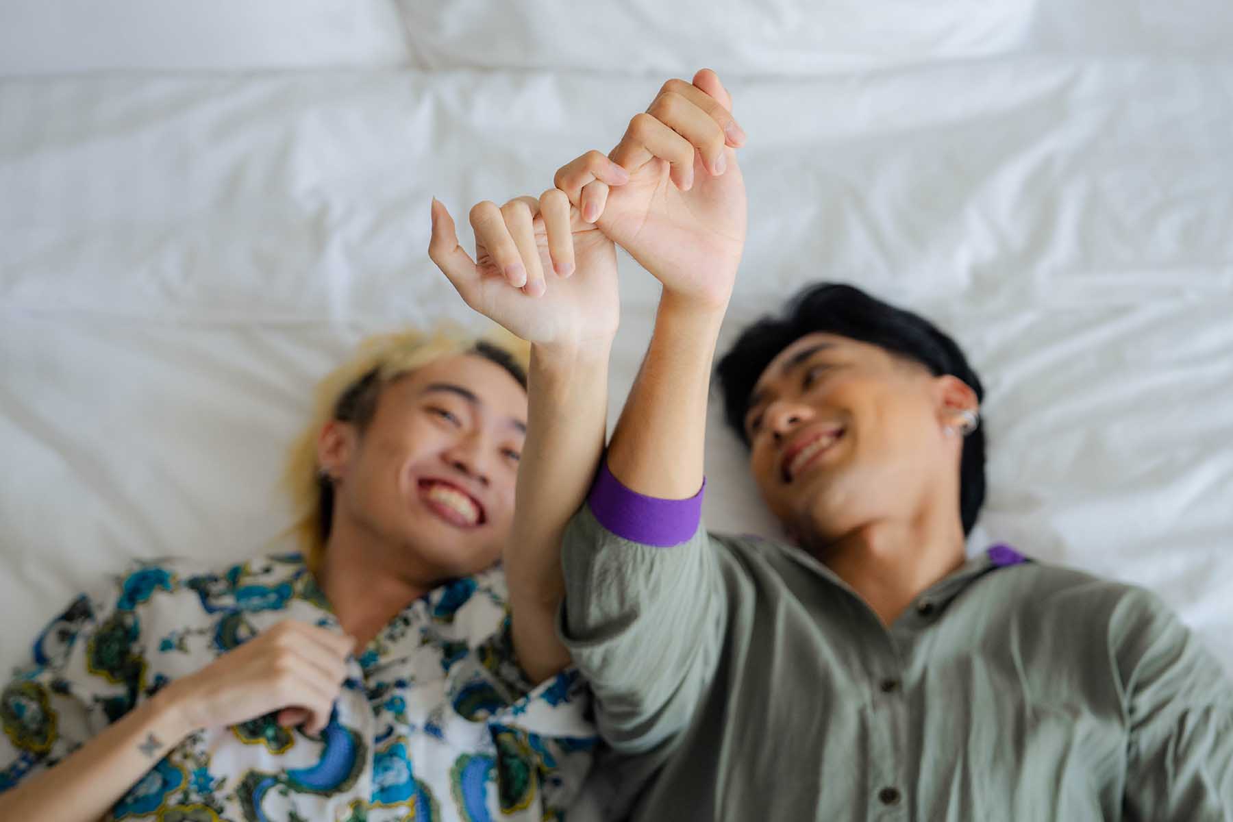 Same-sex couple lying on a bed, smiling. They're holding hands.