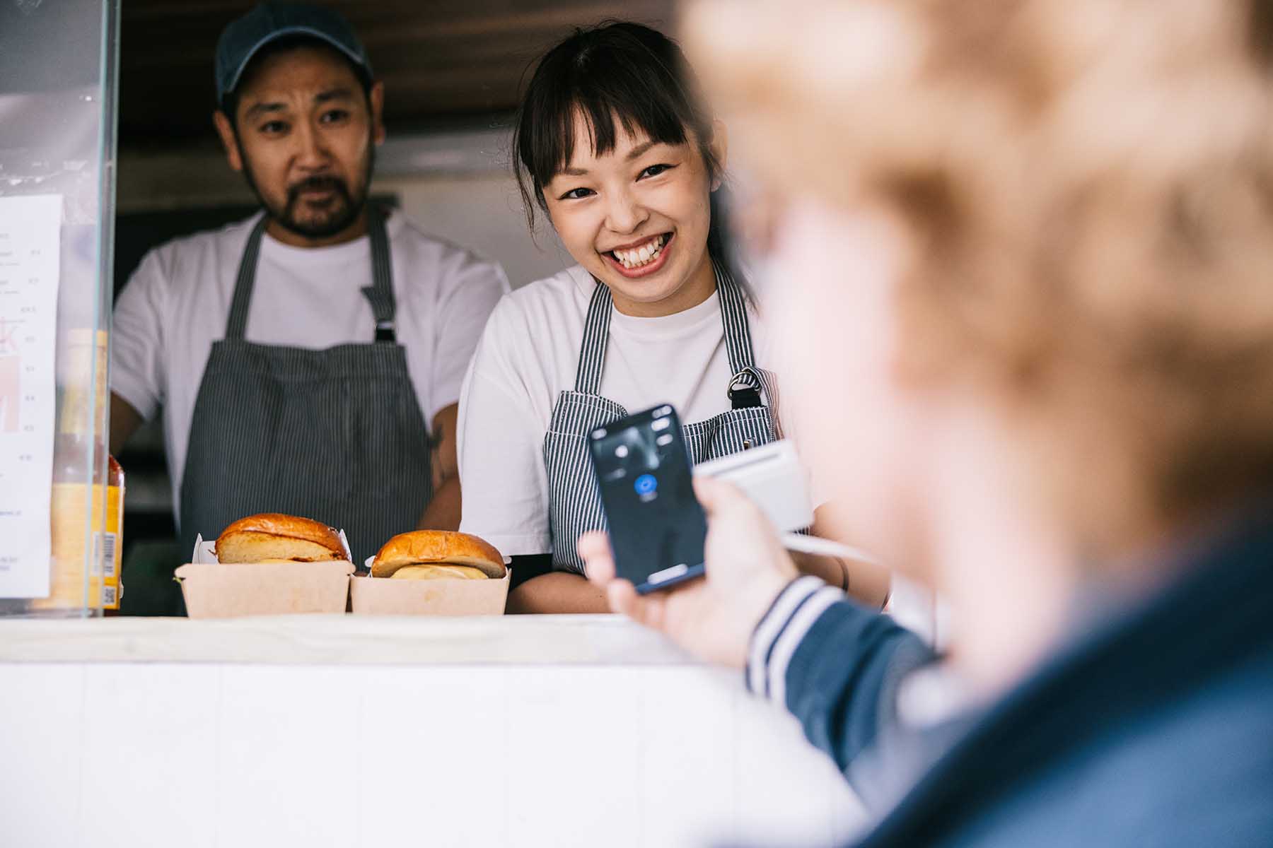 Sales woman of a food truck is smiling as the customer makes an online payment with their phone. Her colleague is standing behind her, looking at the customer somewhat suspiciously.