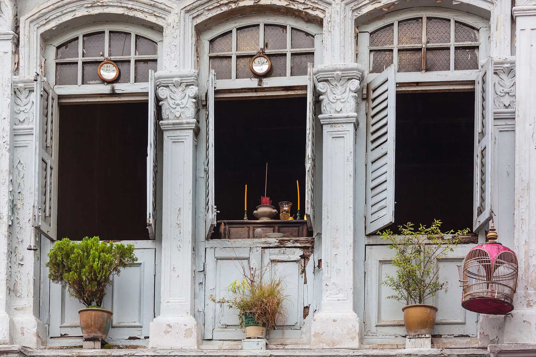 Closeup of an old colonial building facade in historic Little India, Singapore. There are a couple of plants on the ledge and a birdcage is hanging from the sill. The building needs upkeep.