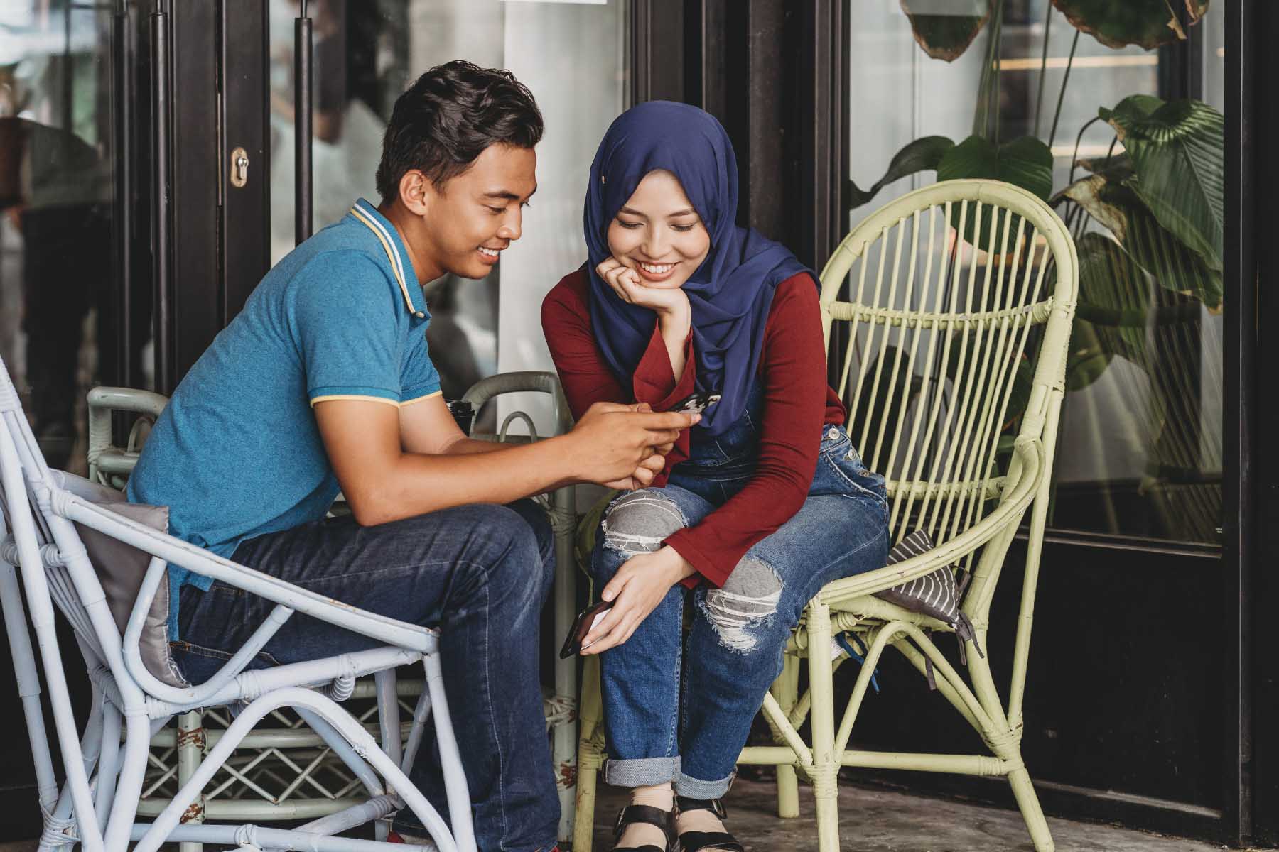 Young couple sitting on chairs at a veranda. Both are smiling like it's puppy love. He's showing her something on his phone.