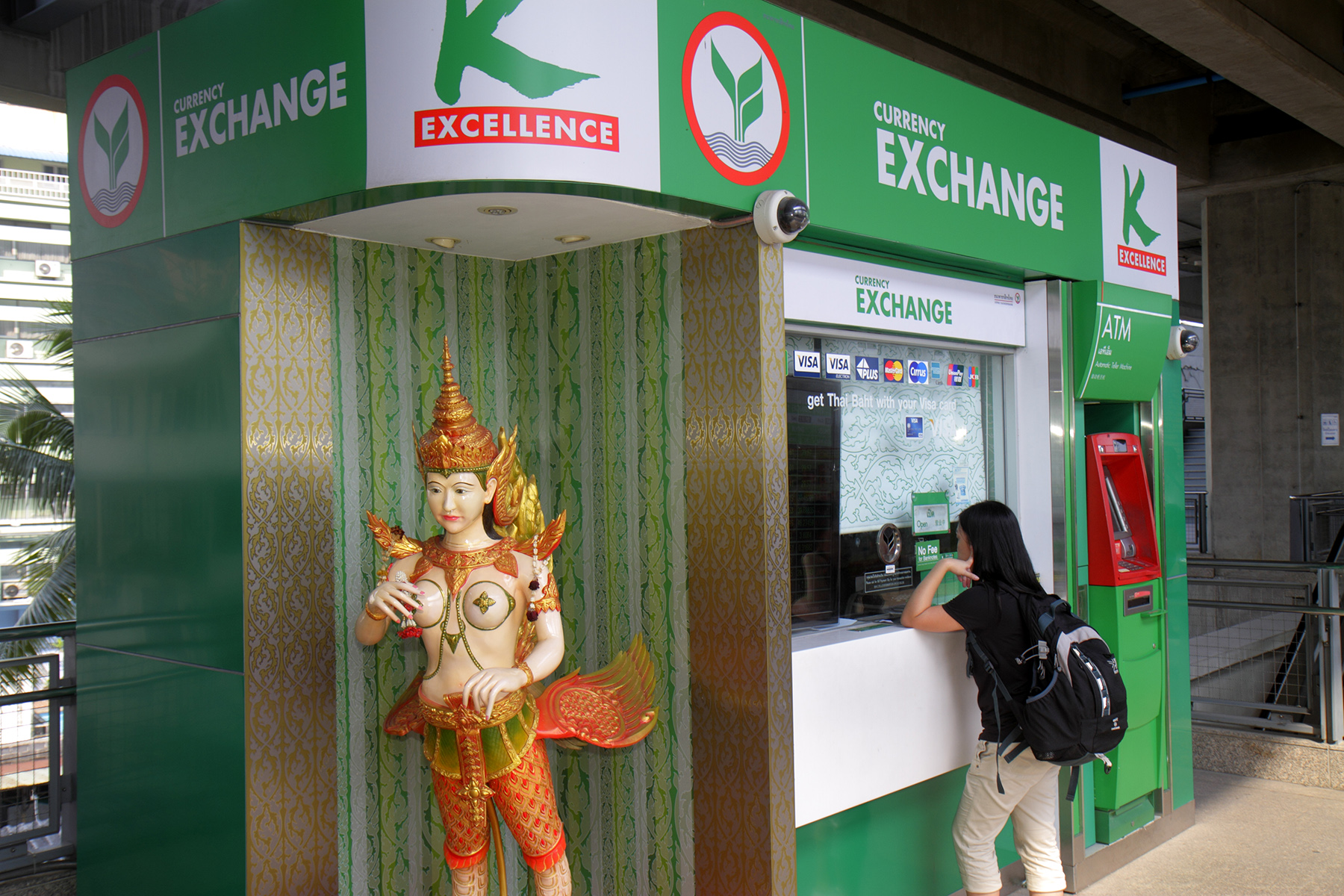 A customer stands at a green currency exchange bank kiosk at Ratchathewi. there is a Thai statue next to it.