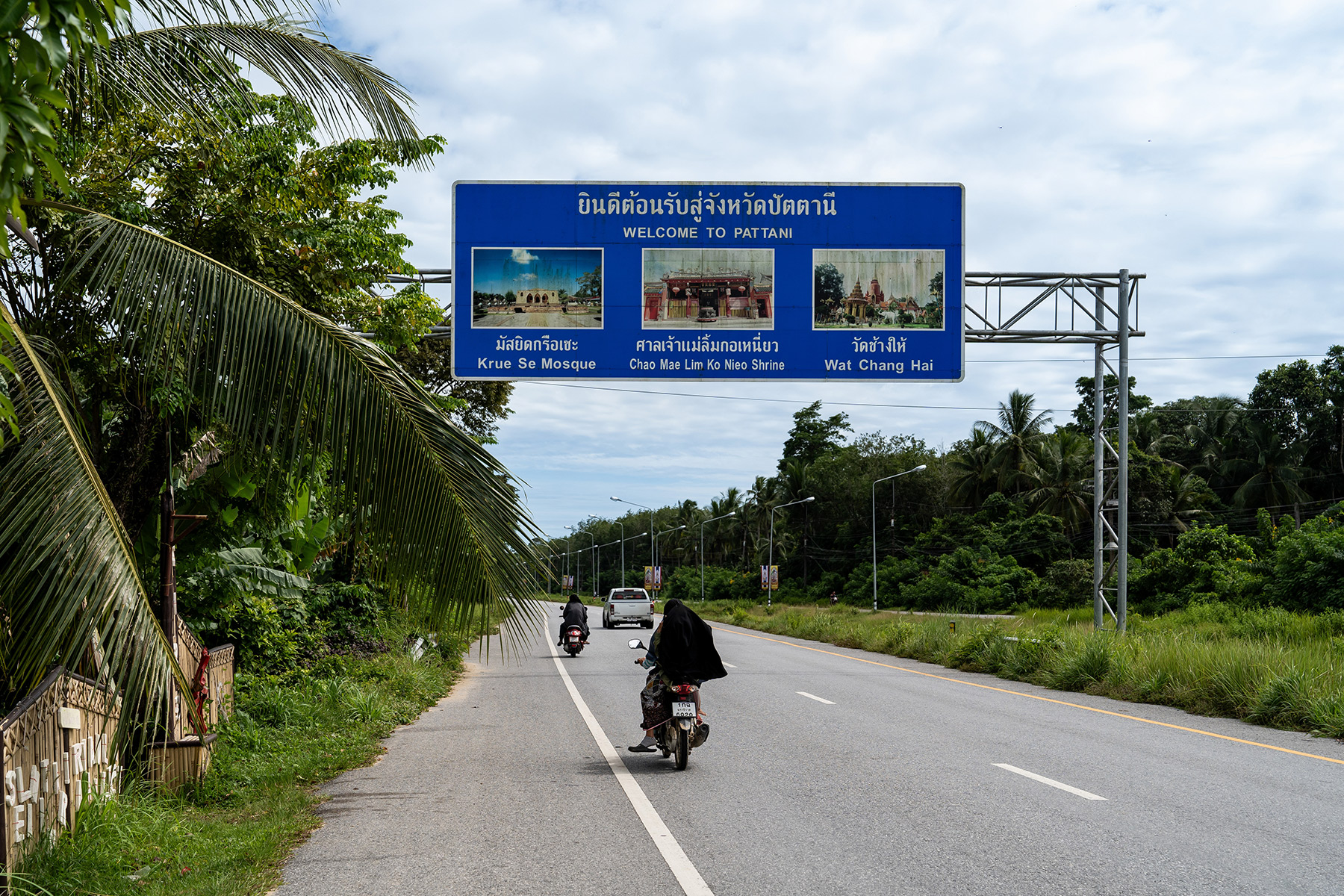 A road sign welcoming motorists to Pattani. People ride motorbikes and drive cars under the sign.