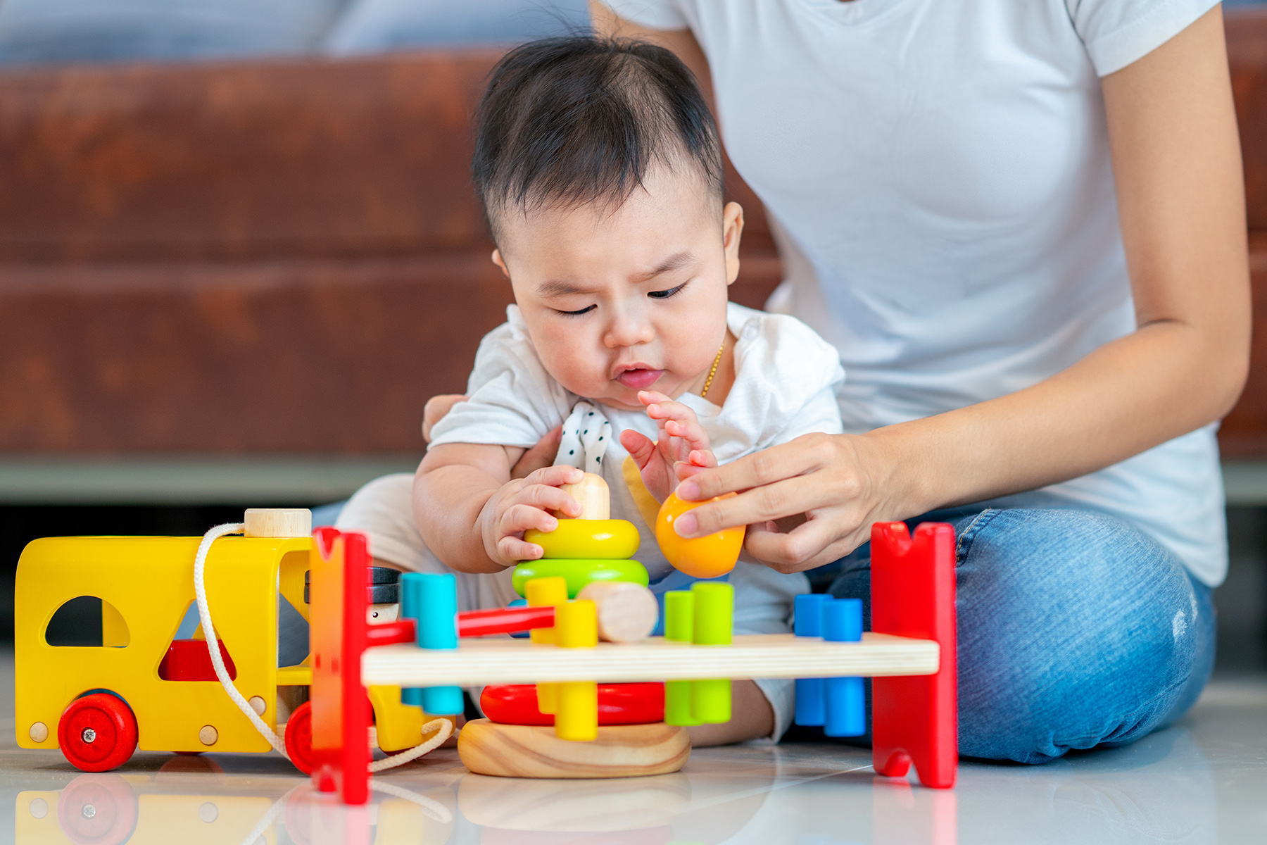 A baby sits on the floor playing with a colorful tool, an adult sits behind them