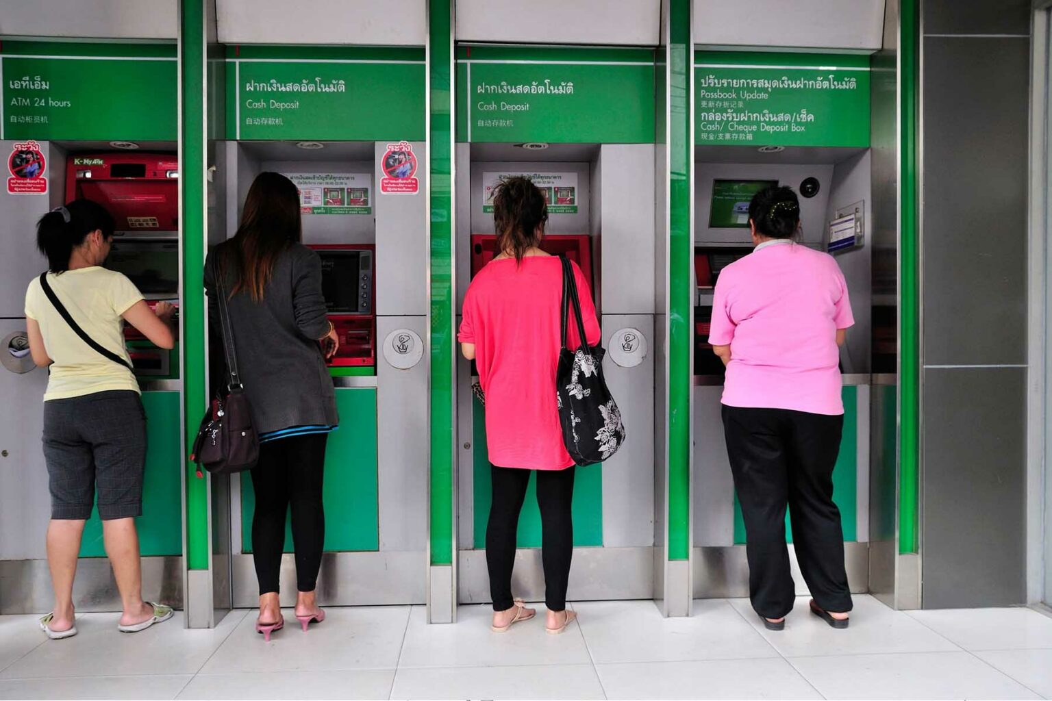 Four women stand in a row drawing money from green ATMs in Thailand.