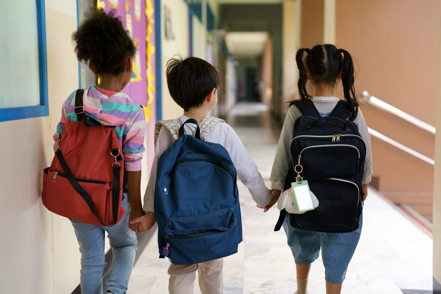Three young children hold hands and walk away from camera down the school hallway. They all wear backpacks and casual clothes