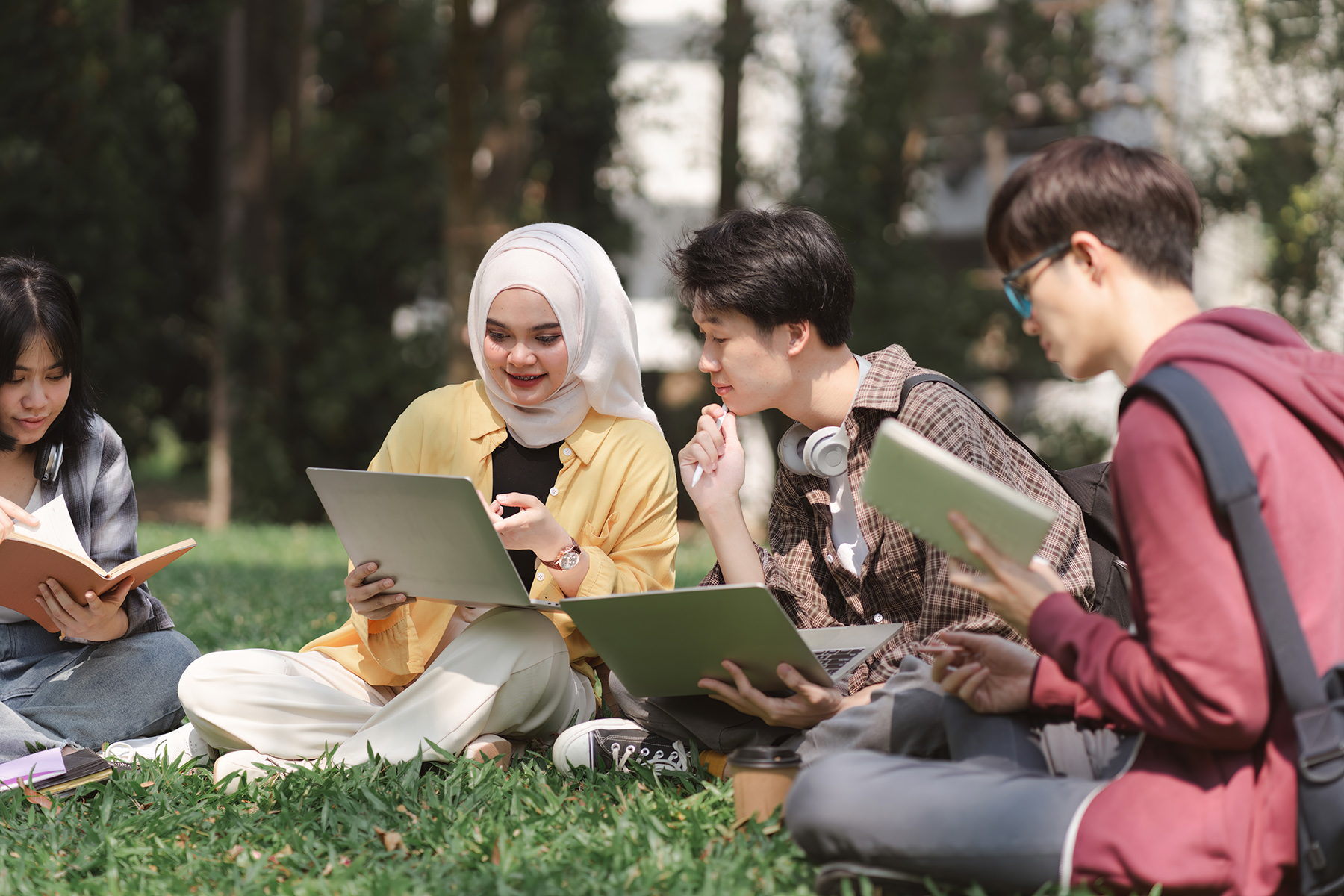 A group of diverse students sits in a circle outside on the grass discussing a project, one holds a laptop