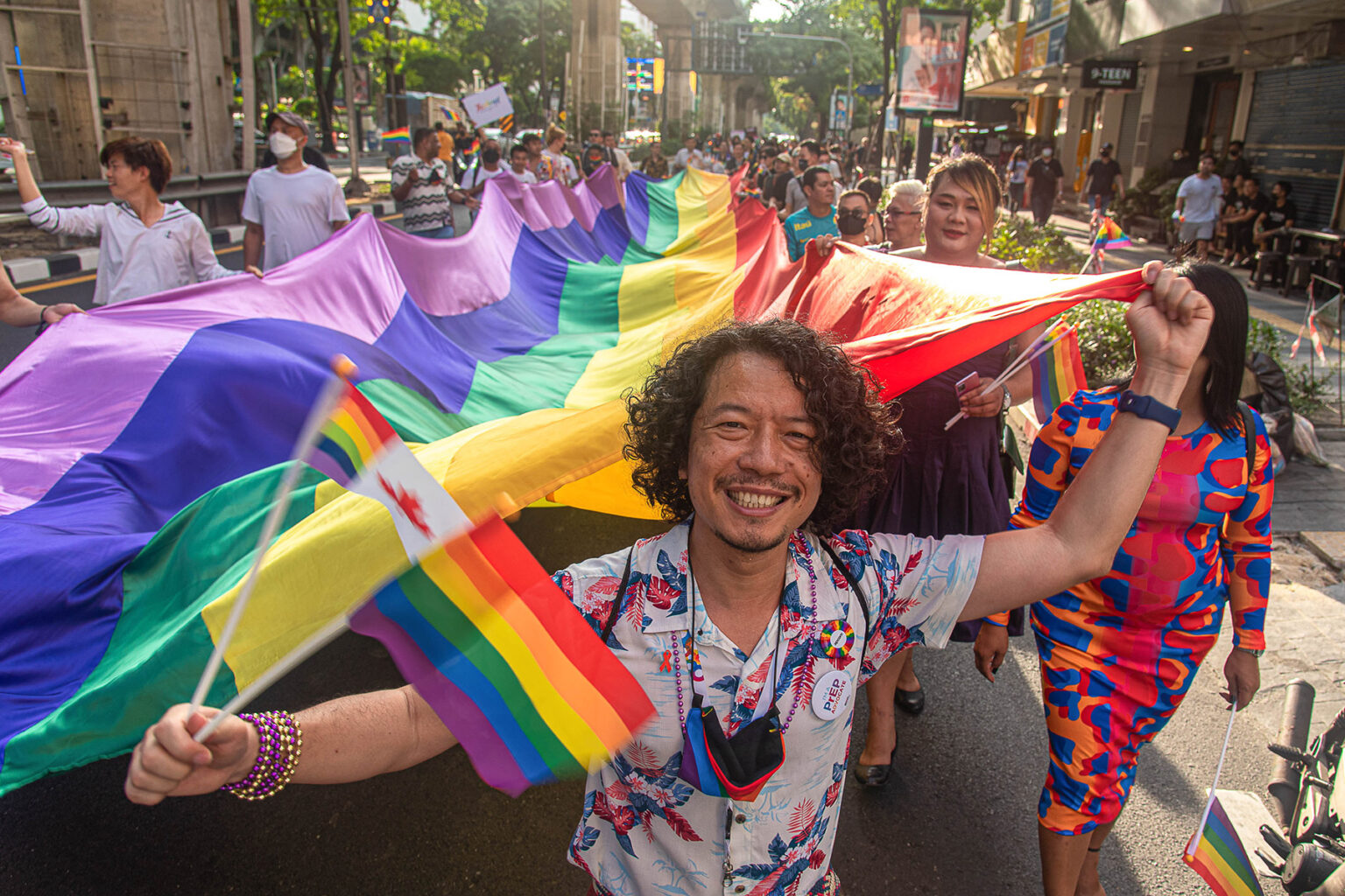 Group of people holding a huge rainbow flag during the Thailand Pride Parade 2022 at Silom road in Bangkok. Man in the front is smiling and holding more small rainbow flags, as well as a Canadian one.