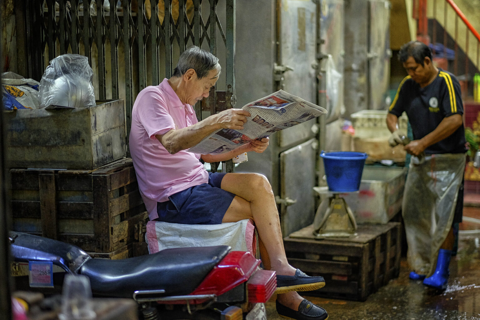 Old man sitting on a crate in a back alley while is reading the newspaper. Someone in the background is cleaning fish before throwing it in a bucket on the scale.