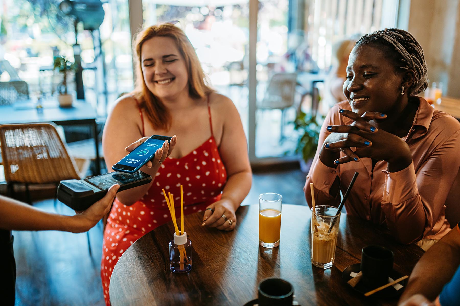 Two smiling friends sitting at a café, drinking juice and coffee. One is paying the bill via a mobile banking application on her phone.