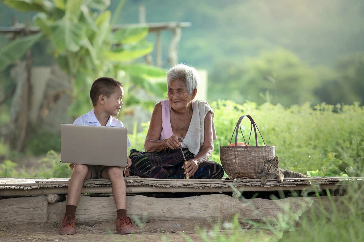 A boy sits next to his grandparent outside, with lots of greenery around, while holding a laptop.