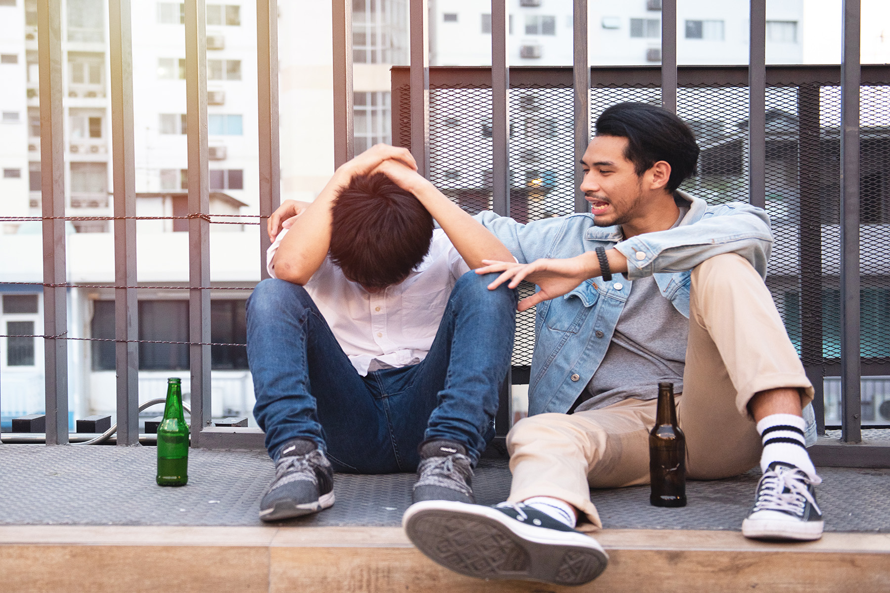 Two guys sitting on the street drinking beer, one has his head in his hands, the other has his arm around his shoulders

