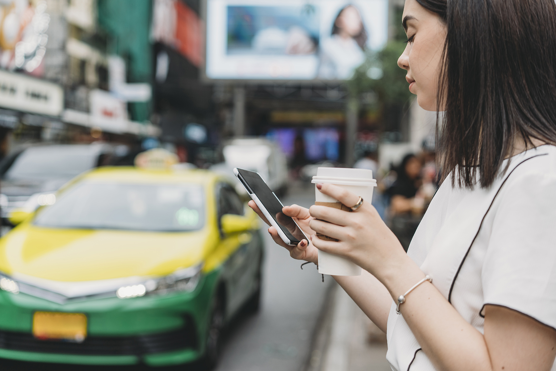 Woman holds a coffee and looks on her mobile app while standing on the street. a green and yellow tax drives in the background