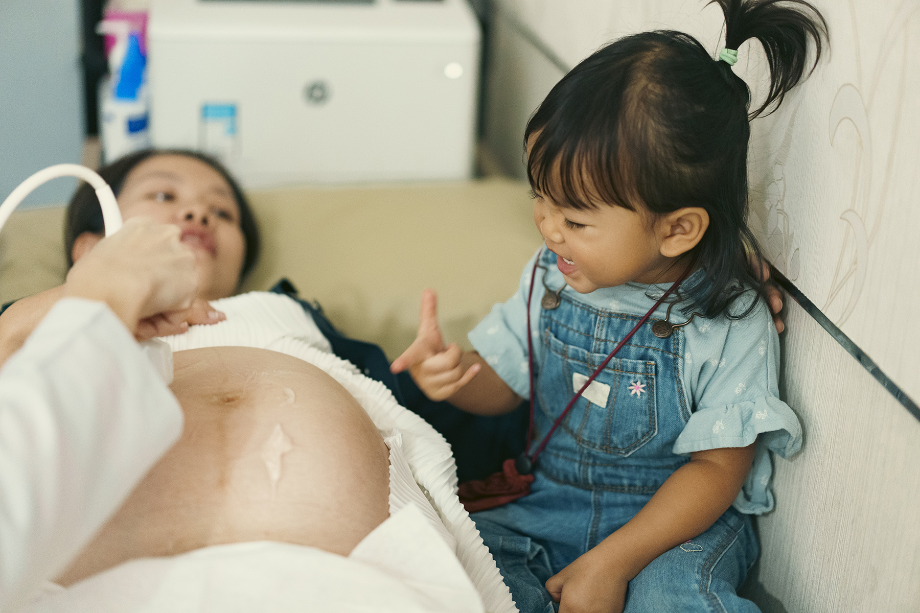 A toddler sits next to her pregnant mom having a ultrasound, she point to her mom's belly