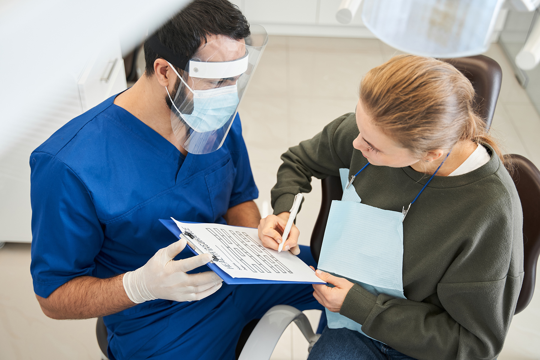 Dental assistant in Thailand holds a clipboard for patient to complete and sign. He wears a full mask and face shield and surgical gloves. She sits in the dentist chair.

