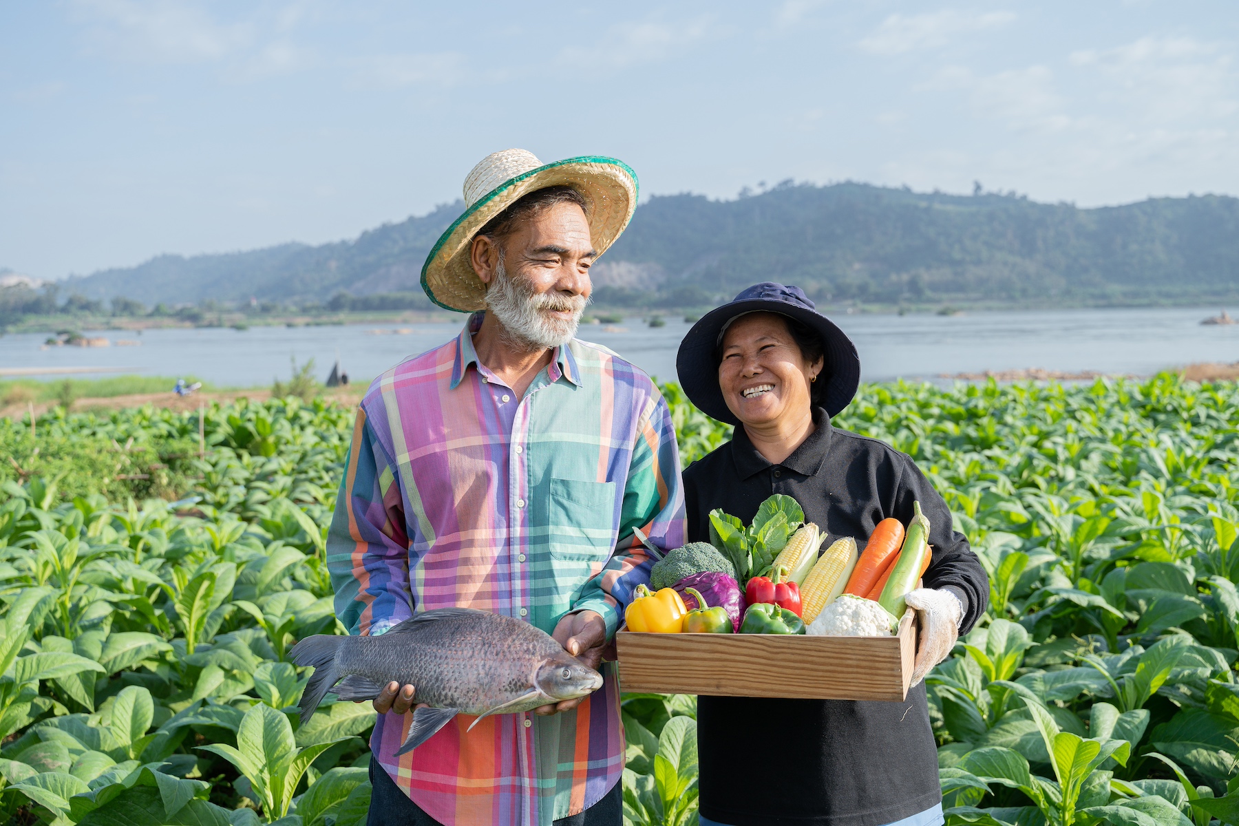 A fisherman and a farmer stand together in a field smiling and holding up their produce