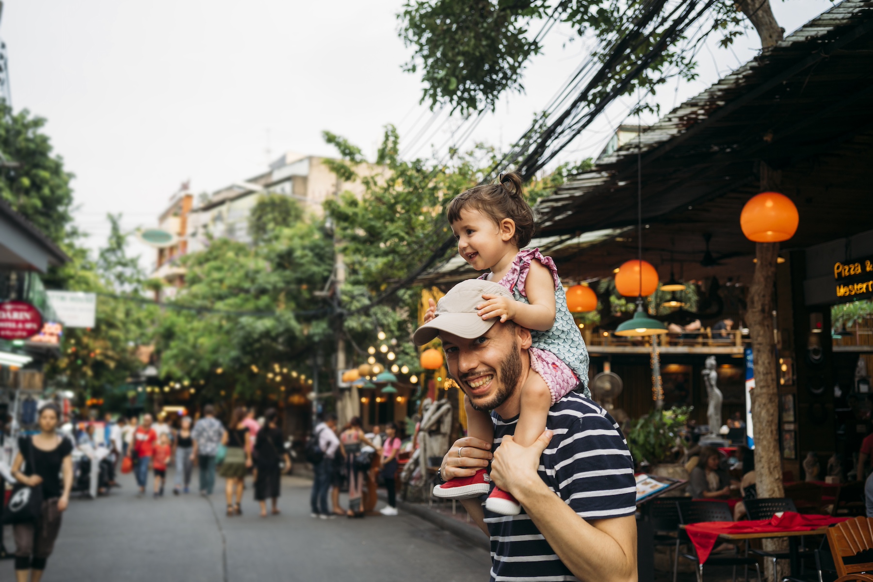 A father smiles holding his young daughter on his shoulders in a crowded Bangkok street
