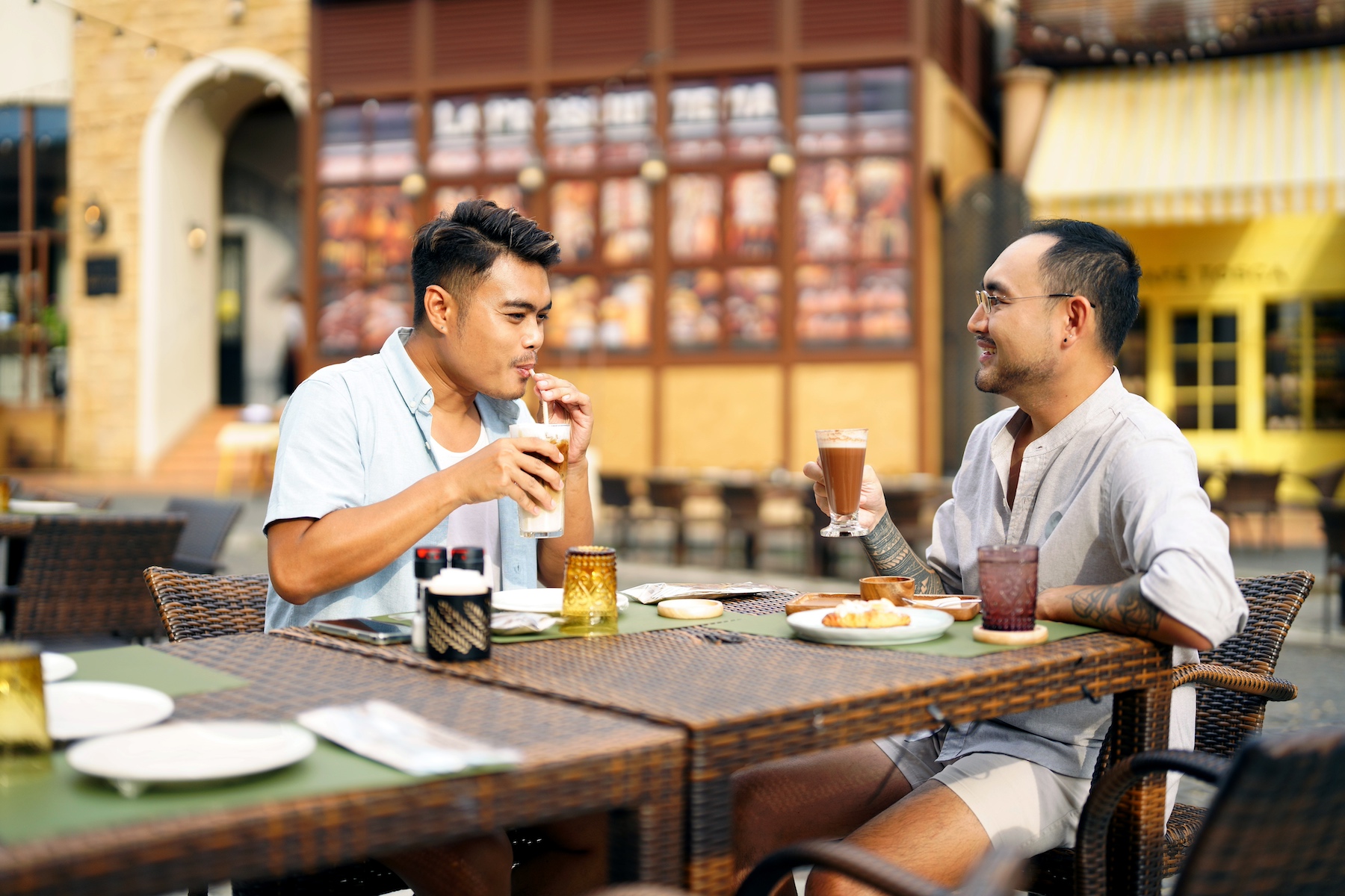 Two men sit at an outdoor cafe with coffee and pastries