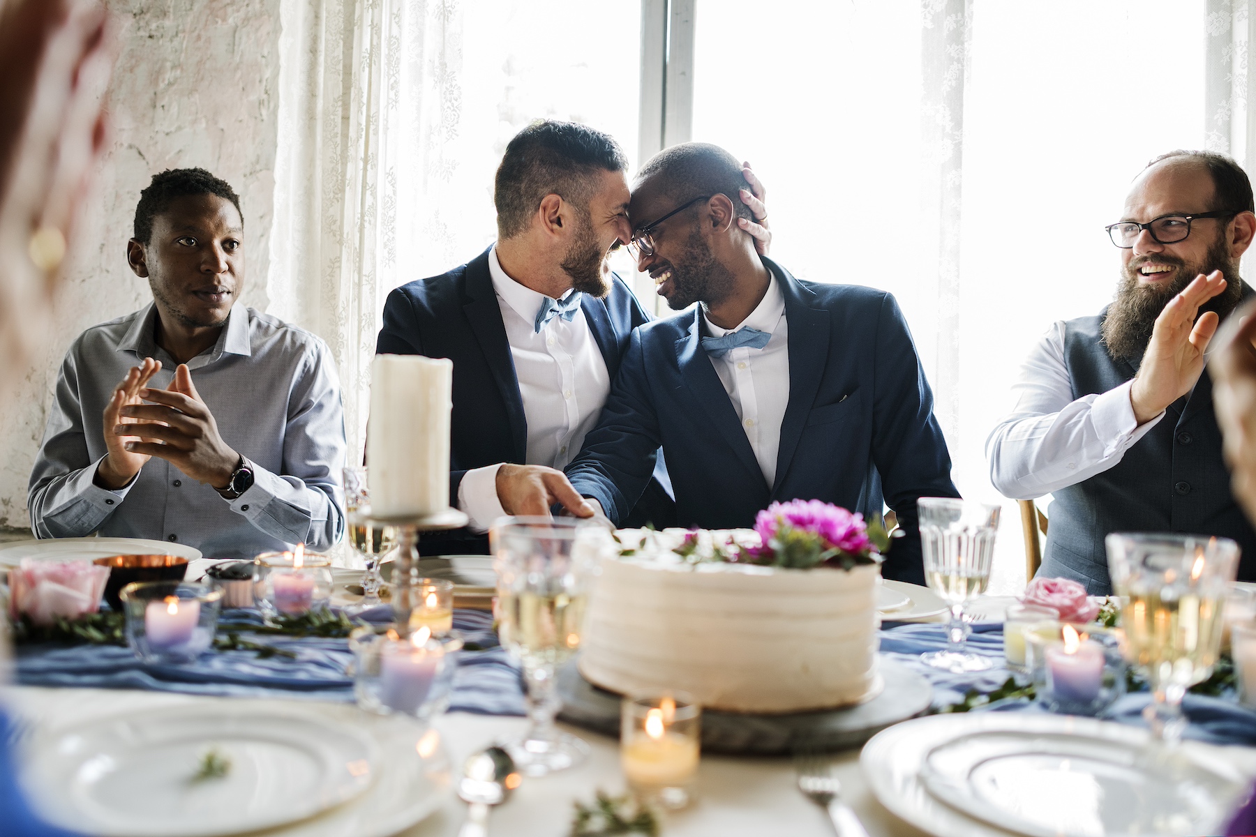 Two grooms sit at a wedding reception table gently cutting the cake and embracing