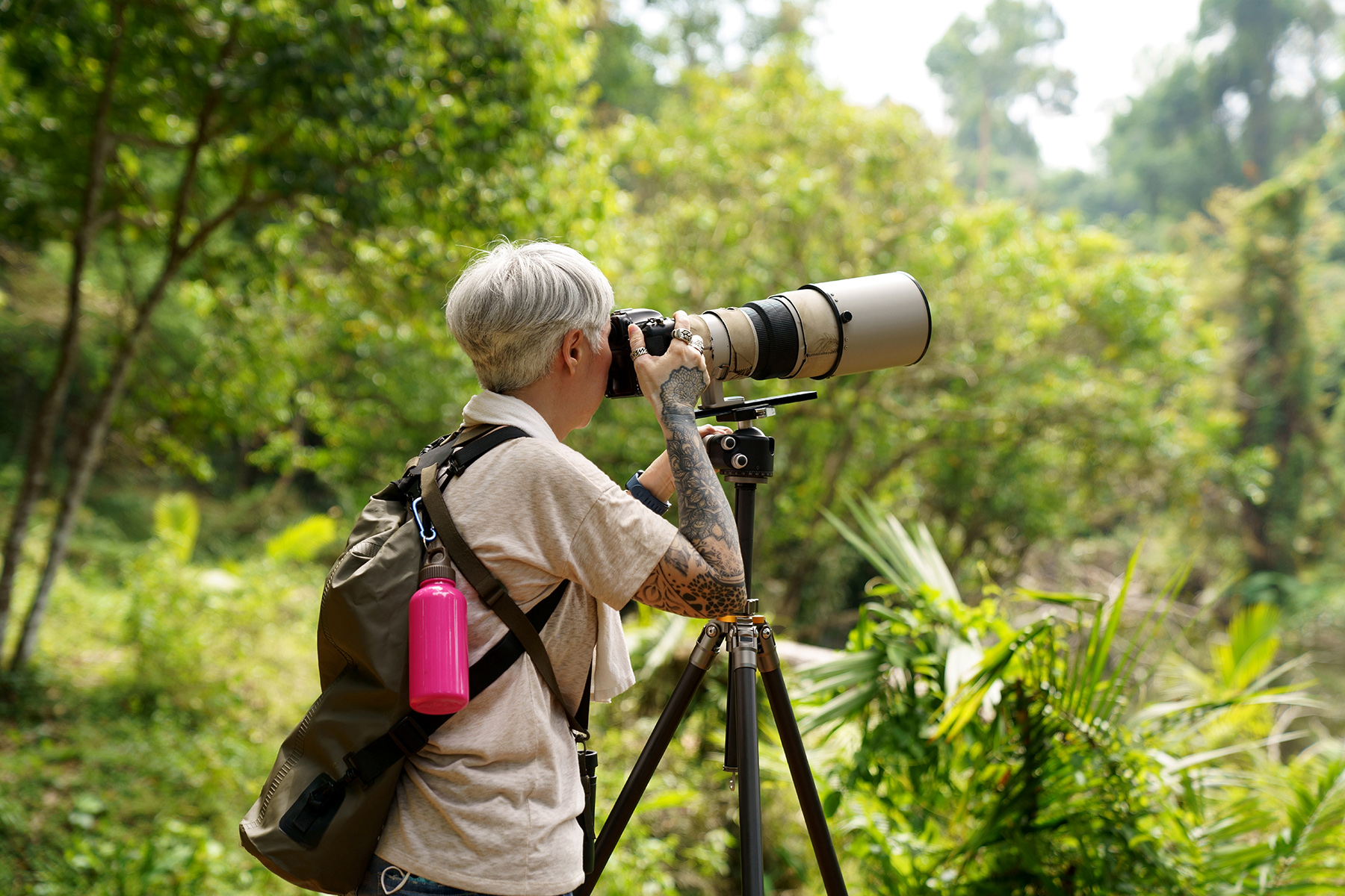 woman with tattoos and a huge camera with a telelens, standing in a forest taking a picture.