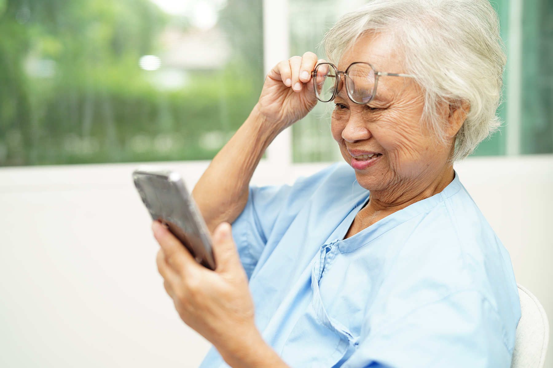 Older woman with grey hair in a hospital gown lifts her glasses to read on her phone