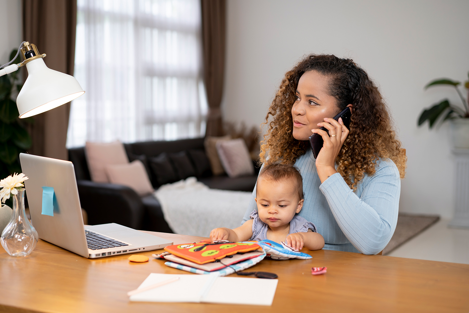 Woman working on a laptop while taking a phone call. A baby sits on her lap.