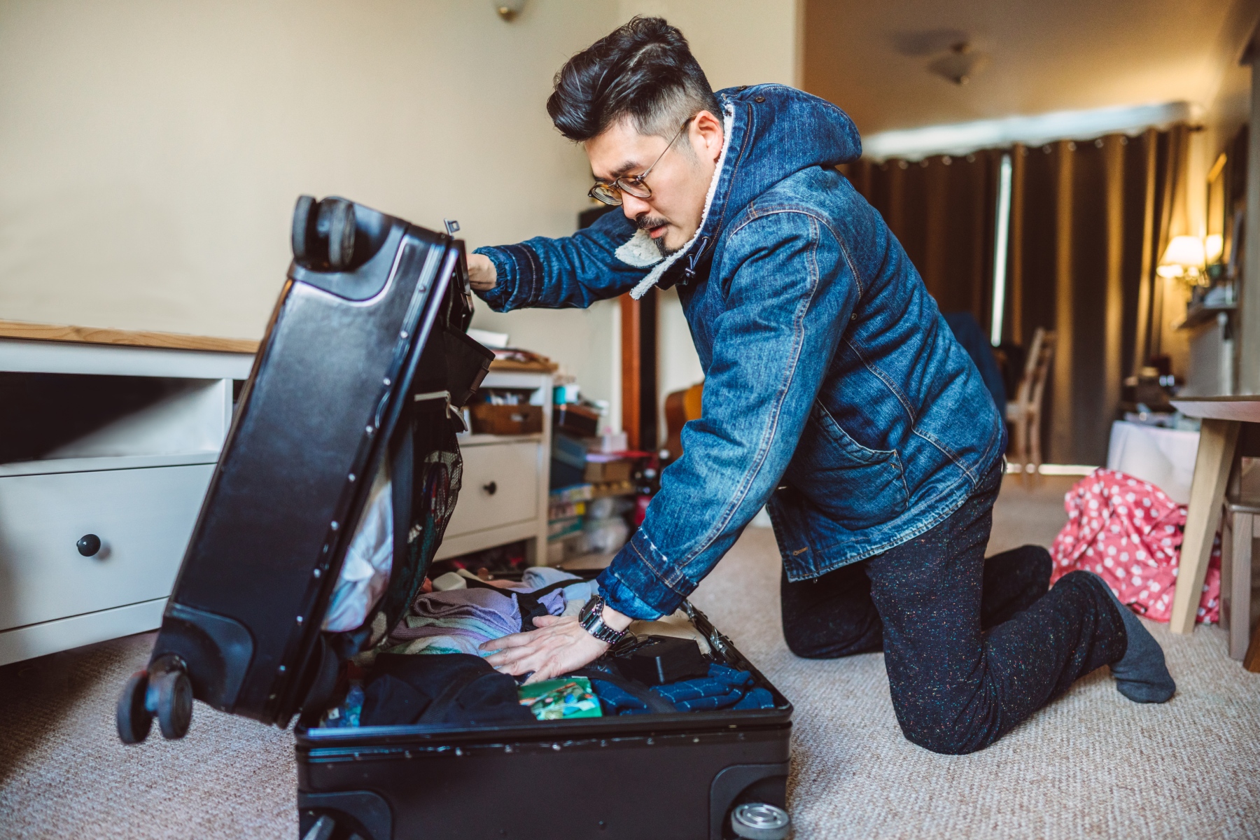 Man wearing denim jacket packs large suitcase in his room