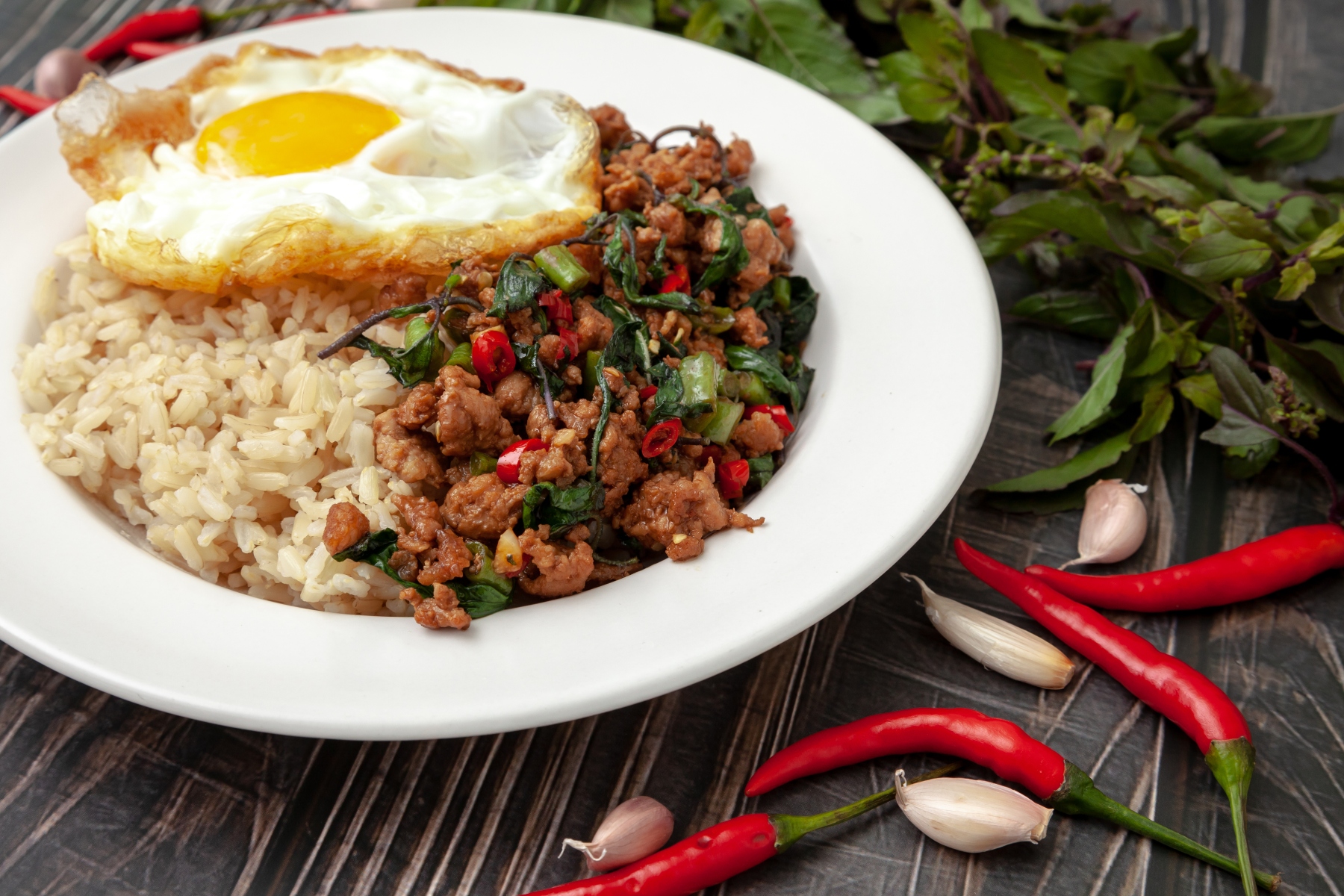Pad kra pao moo served with a fried egg on top, in a plate alongside chillies and garlic cloves