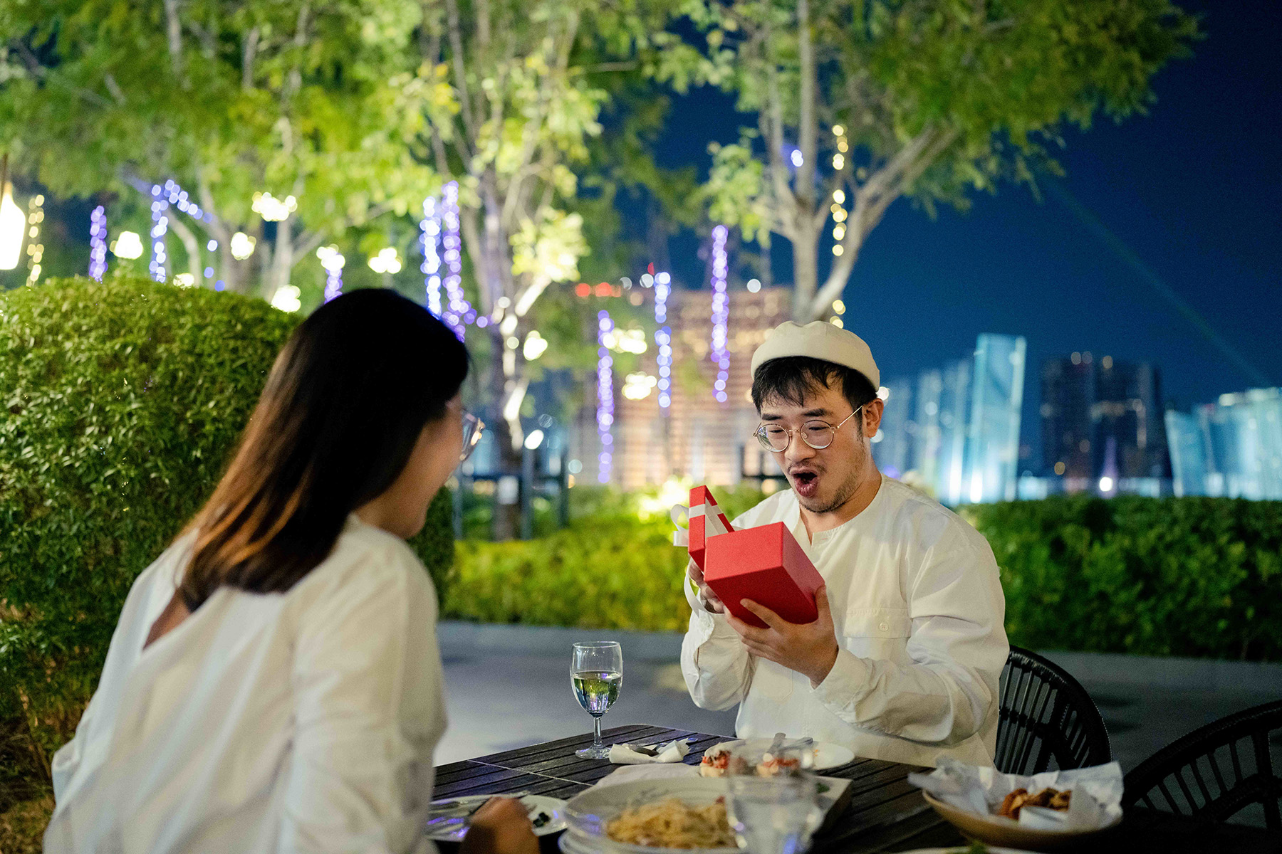 A young guy smiles surprised as he opens a gift in a red box while sitting in a restaurant with his girlfriend

