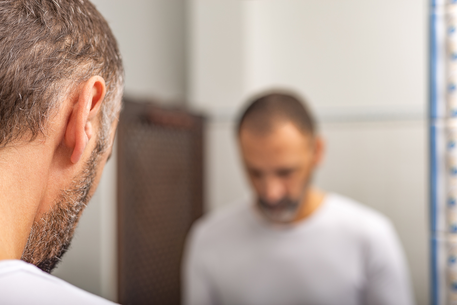 Blurry reflection in bathroom mirror of a man with a short hair and a stubble beard in whirt shirt looking down, looking worried