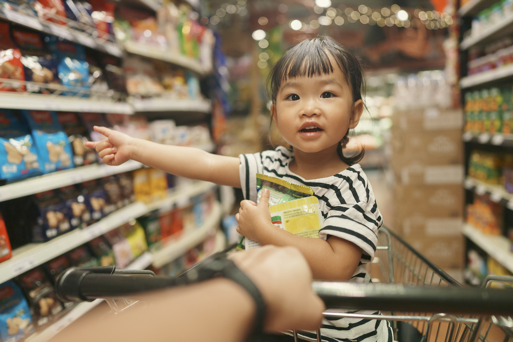 Little girl holds a packet of sweets and points to something in the shop aisle  while sitting in the shopping cart

