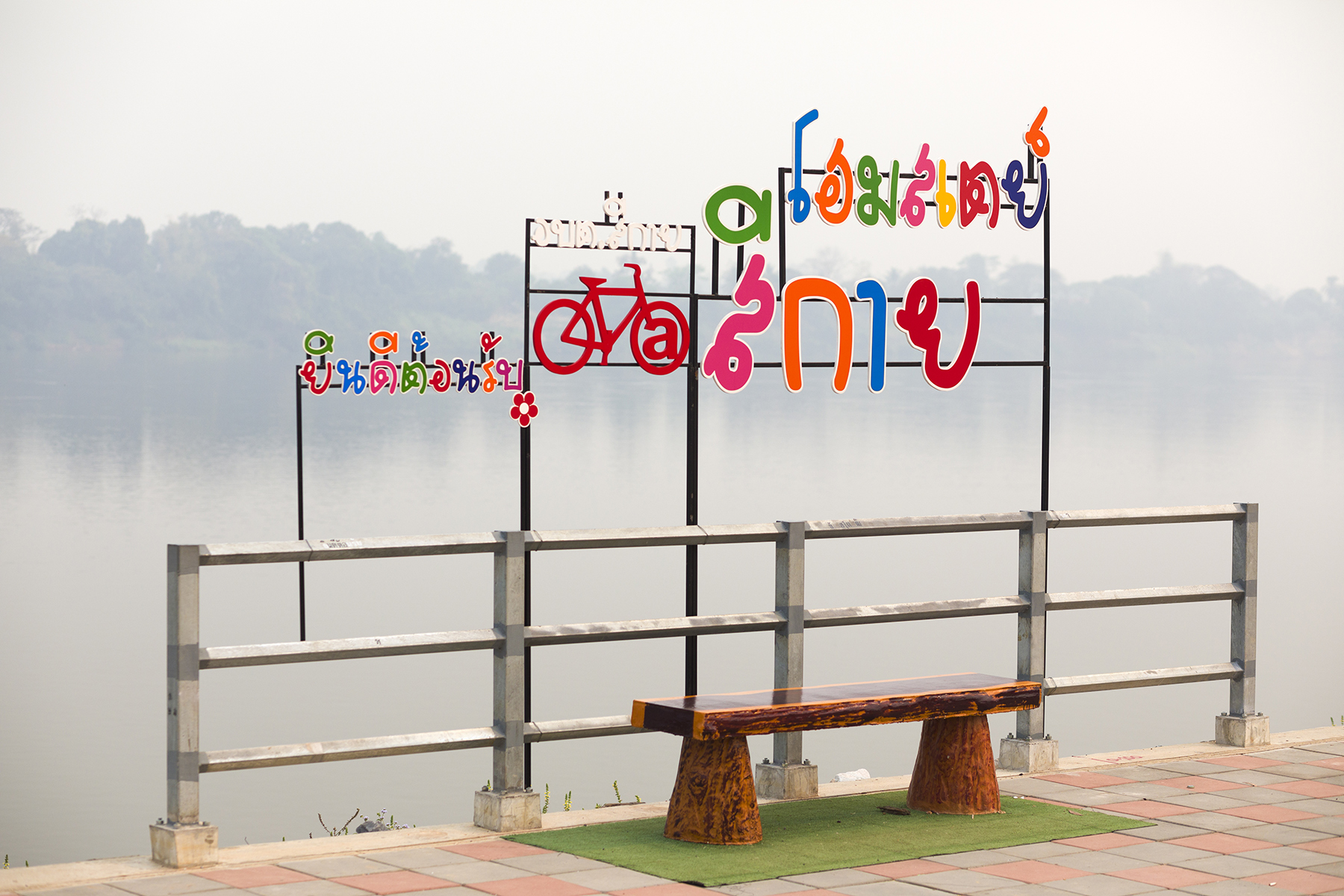 Brightly colored Thai sign with a bicycle on it. The sign is by a river.