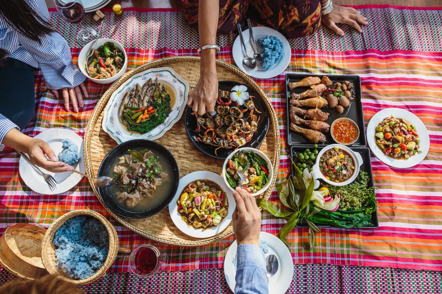 A spread of various Thai dishes on a mat, with various hands reaching out to serve the food