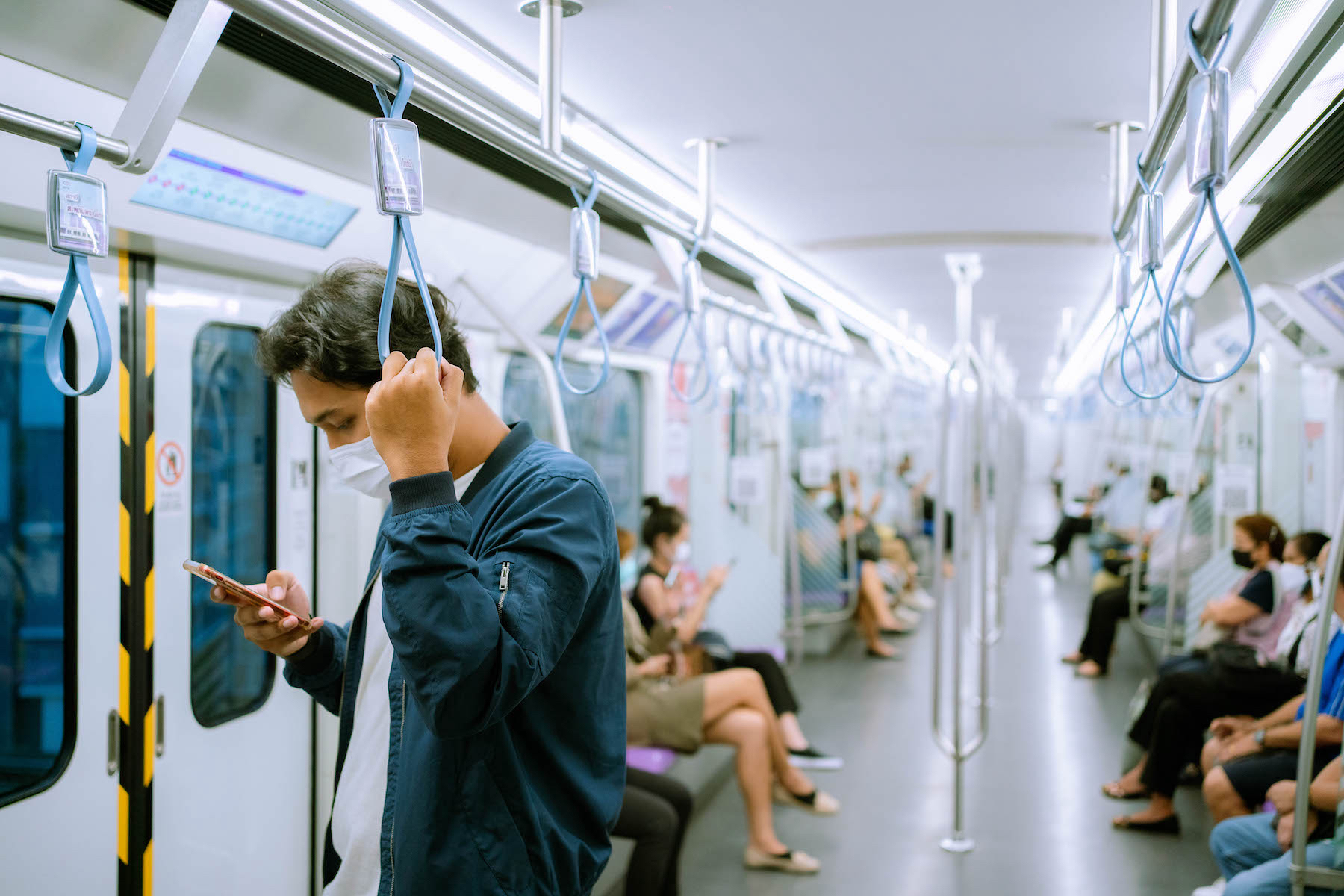 A man stands on a Thai subway train wearing a protective face mask and looking down at his phone
