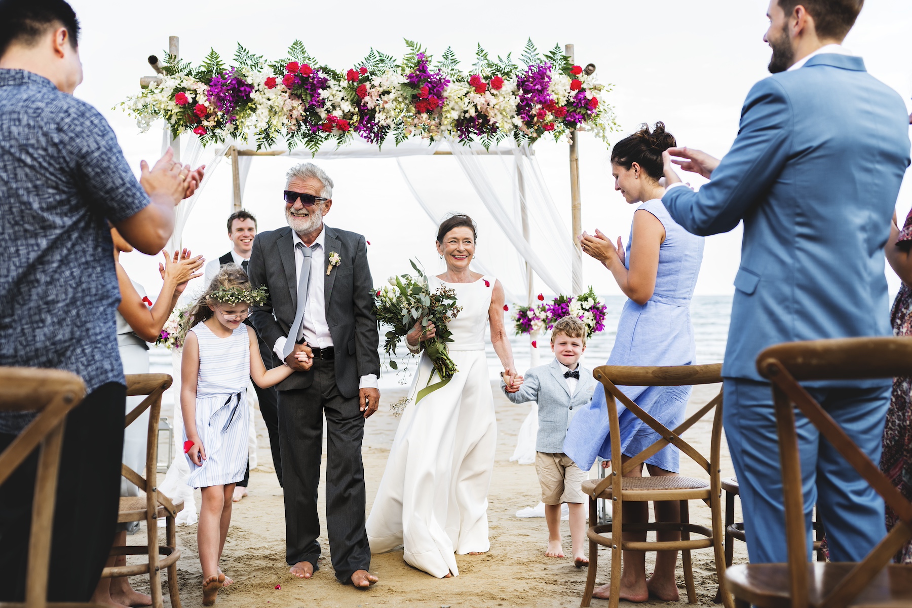 A newly married older couple walks down the aisle at a beach wedding holding their grandchildren's hands